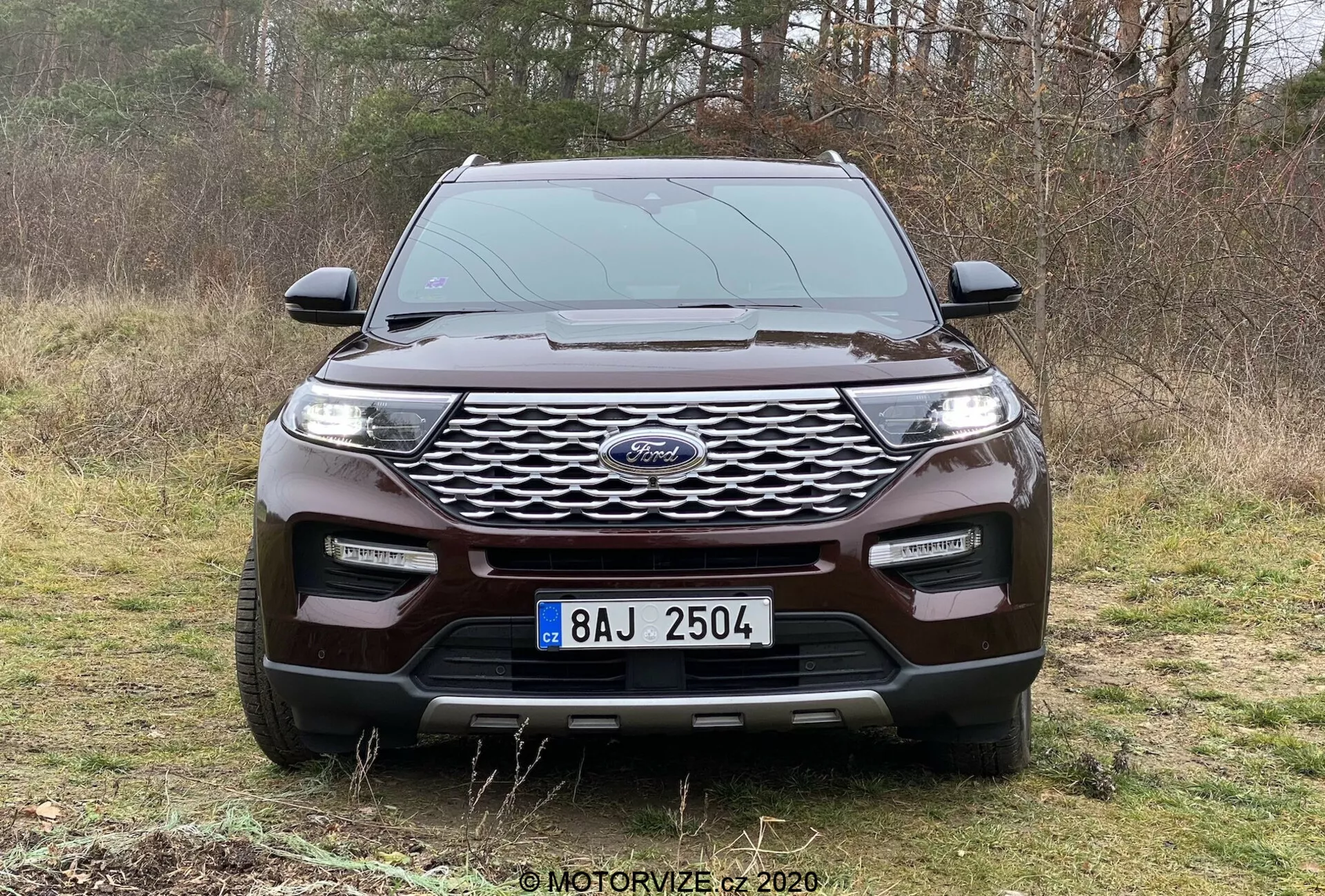 Sprednji pogled na SUV Ford Explorer 2019, parkiran na travmati površini z drevesi v ozadju, prikazuje zunanje značilnosti moderne vozila. Izrazita rešetka s centralnim emblemom, elegantne LED prednje luči, robusten sprednji odbijač z meglenkami, srebrni element za robusten videz in vidne aerodinamične linije pokrova motorja. Primerno za razprave o avtomobilskem oblikovanju, varnostni tehnologiji in zmogljivostih za vožnjo po terenu.