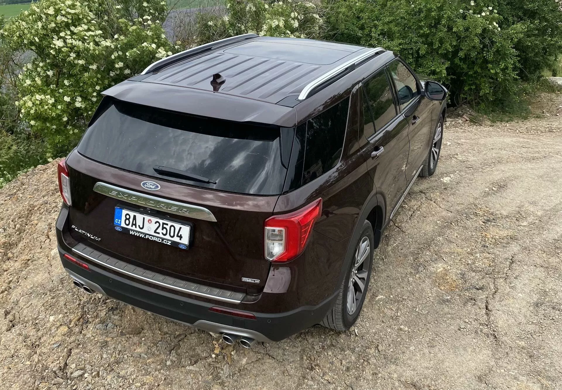 Dvignjen zadnji trikraten kotni pogled na temno obarvani Ford Explorer 2019, ki poudarja zadnji del vozila in bočni profil. Vidne značilnosti vključujejo logotip Ford, ime modela 'Explorer', edinstvene zadnje luči, brisalec zadnjega vetrobrana, anteno v obliki plavuti morskega psa, dvojni izpušni sistem, vzvratno kamero, zlitinska kolesa, zatemnjena okna in stranska ogledala z vgrajenimi smerniki. SUV je nekoliko desno izven središča na neutrjenem terenu, kar nakazuje lahkotno terensko zmogljivost.