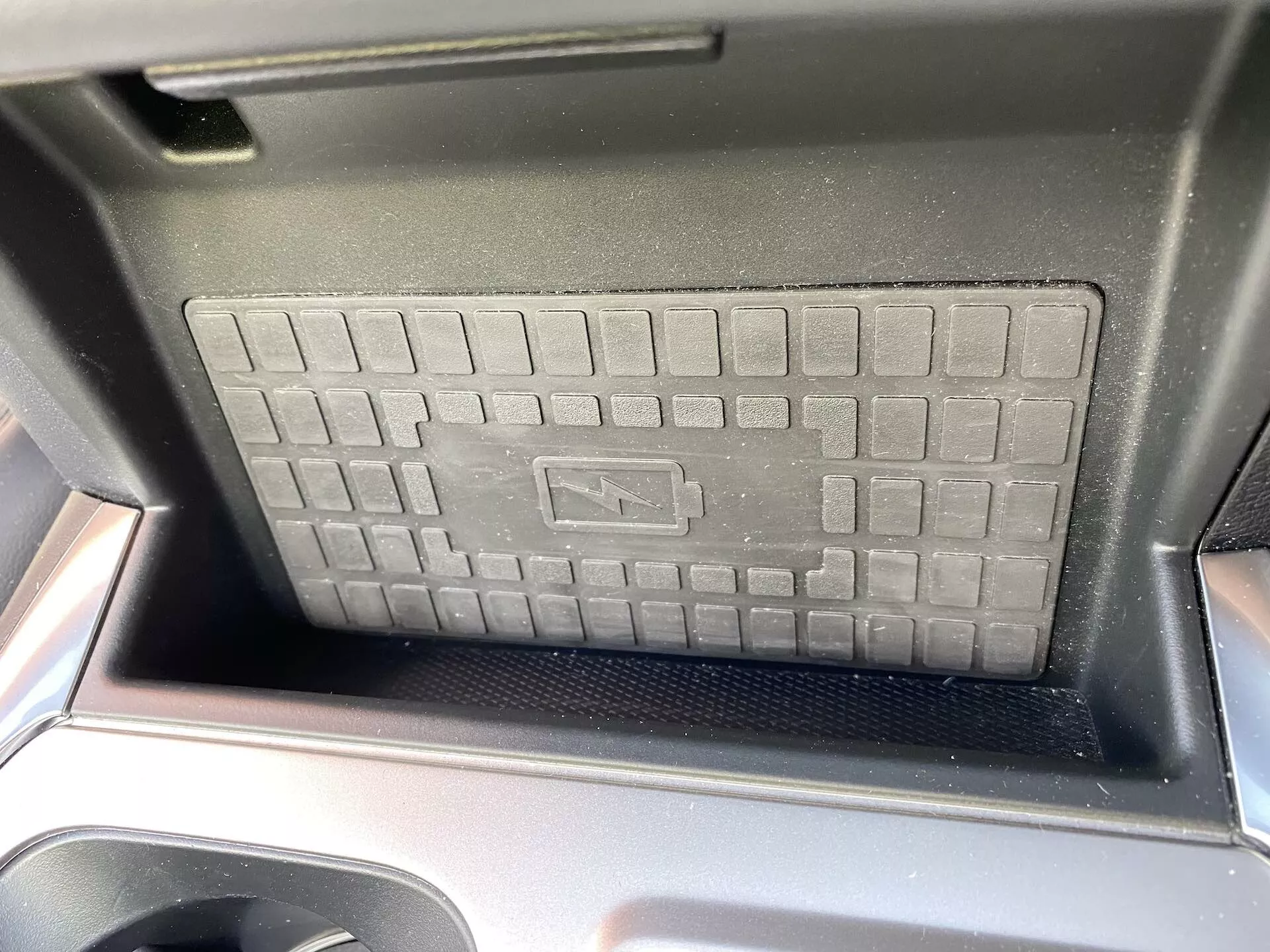 Podroben pogled na predel osrednje konzole Ford Explorer 2019, ki poudarja brezžično polnilno ploščico z dvignjeno, mrežasto teksturo in nedrsečo površino. Vidni so sijoči črni plastični okviri, ki dodajo pridih sodobnega avtomobilskega dizajna. Sončna svetloba kaže na dnevni fotografiji iz sprednjega sedeža, simbol polnjenja pa označuje napredno brezžično polnilno tehnologijo.