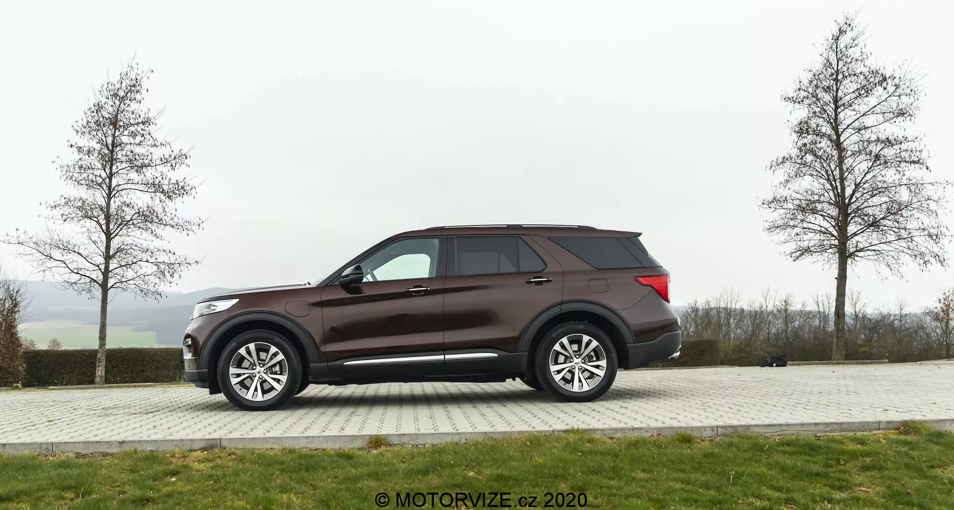 Stranski profil SUV-ja Ford Explorer 2019, parkiranega na tlakovanem območju s travo v ozadju na oblačen dan. Vozilo ima velika večkraka lita platišča, zatemnjena okna in temno rjavo ali bordo lakiranje. Oblikovne lastnosti vključujejo stranska ogledala verjetno z električnim nastavljanjem in integracijo smernikov, aerodinamično linijo strehe in vidno zadnjo anteno.