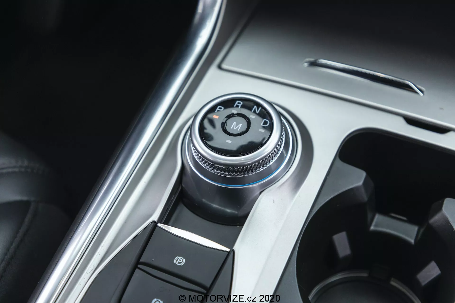 Pogled od blizu na gumb za izbiro prestav in del sredinske konzole v notranjosti avtomobila Ford Explorer 2019. Osrednji del je rotacijski selektor prestav z oznakami 'P, R, N, D', ki označujejo avtomatski menjalni sistem. Fotografija je posneta iz rahlo premaknjenega, zgornjega kota. Okoli selektorja prestav so gumbi, vključno z enim s simbolom parkirne zavore, kar nakazuje na sistem elektronske parkirne zavore. Konzola združuje kovinske in plastične površine, poudarjajoč vrhunskost notranjosti vozila. Dobro osvetljena slika poudarja kovinske elemente, z majhno globinsko ostrino osredotočeno na selektor prestav.