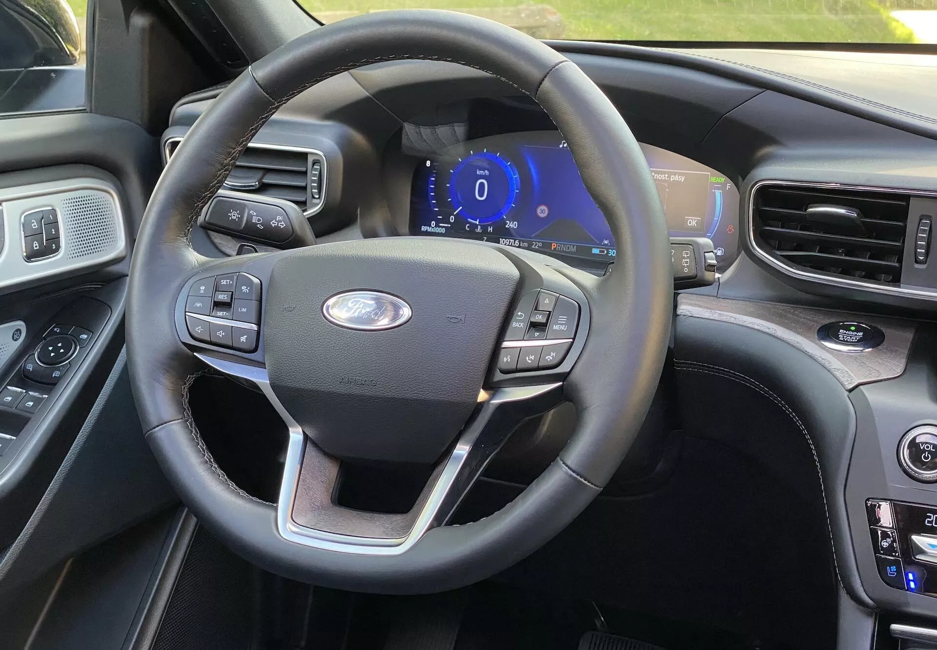 Pogled na notranjost Ford Explorer 2019 iz voznikovega sedeža, ki prikazuje trikraki volan z večfunkcijskimi gumbi za tempomat, nastavitve zvoka in povezljivost telefona. Sredinsko nameščen Fordov emblem in digitalna armaturna plošča osvetljena z modro barvo nakazujeta, da je vozilo v načinu dodatne opreme ali zagona. Na desni je videti del osrednje konzole z gumbi za klimatske nastavitve in nastavitvami temperature, kar kaže na dvojno klimatsko napravo. Notranjost vsebuje mešanico usnjenih in lesnih oblog za premium občutek, z drugim večjim zaslonom, ki je rahlo viden na robu.