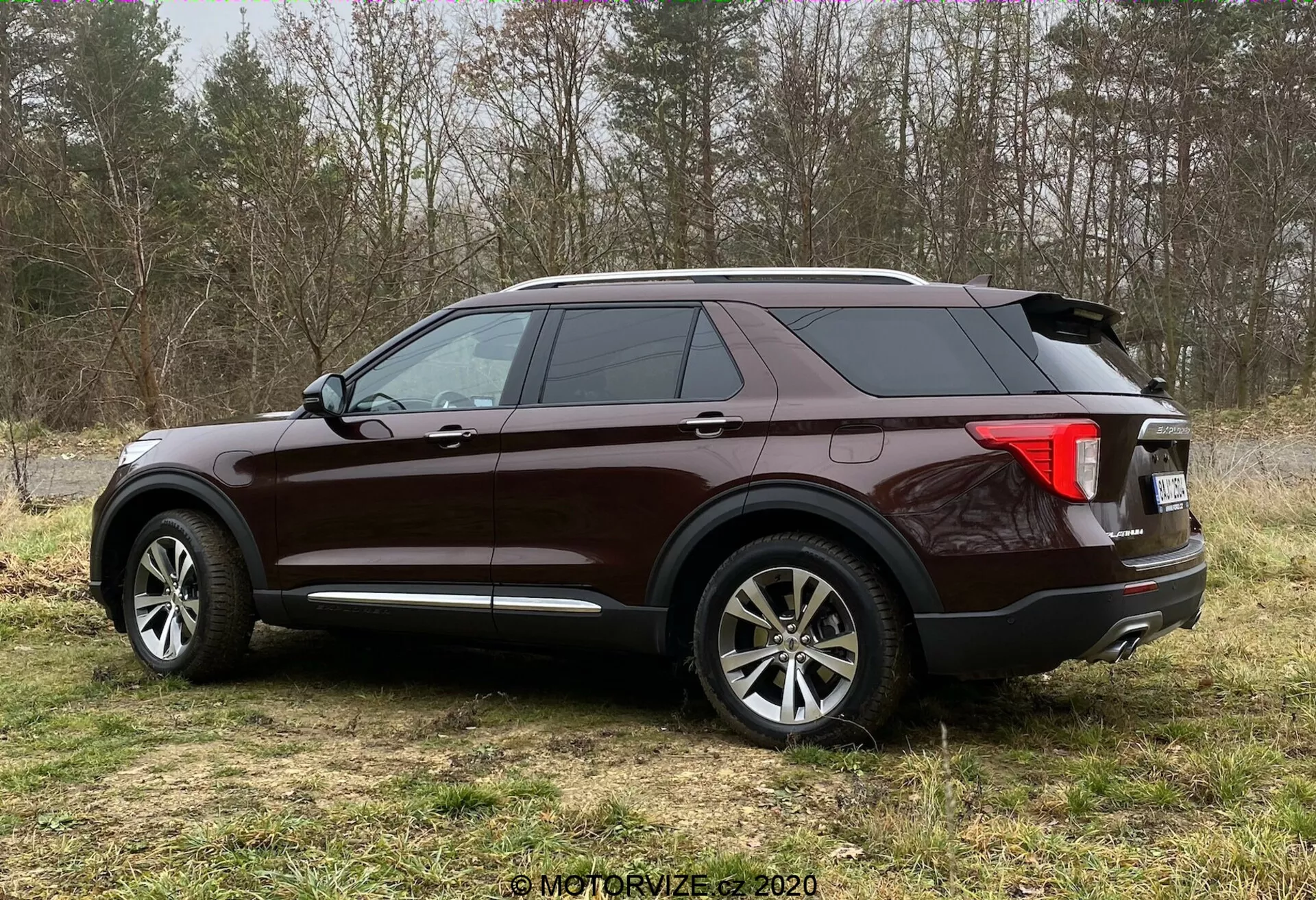 Stranski pogled na SUV Ford Explorer 2019 v bogatem bleščečem bordo laku, postavljen na travnato površino s pol-gozdnim ozadjem. Trikotni kot prikazuje velika večkraka aluminijasta platišča, padajočo zadnjo strešno linijo, zatemnjena okna, izrazito bočno značilno črto, zadnje luči, ki obkrožajo vozilo, verjetno z LED tehnologijo, antena morski pes, kromirane dodatke, strešne nosilce in dvojni izpušni sistem.