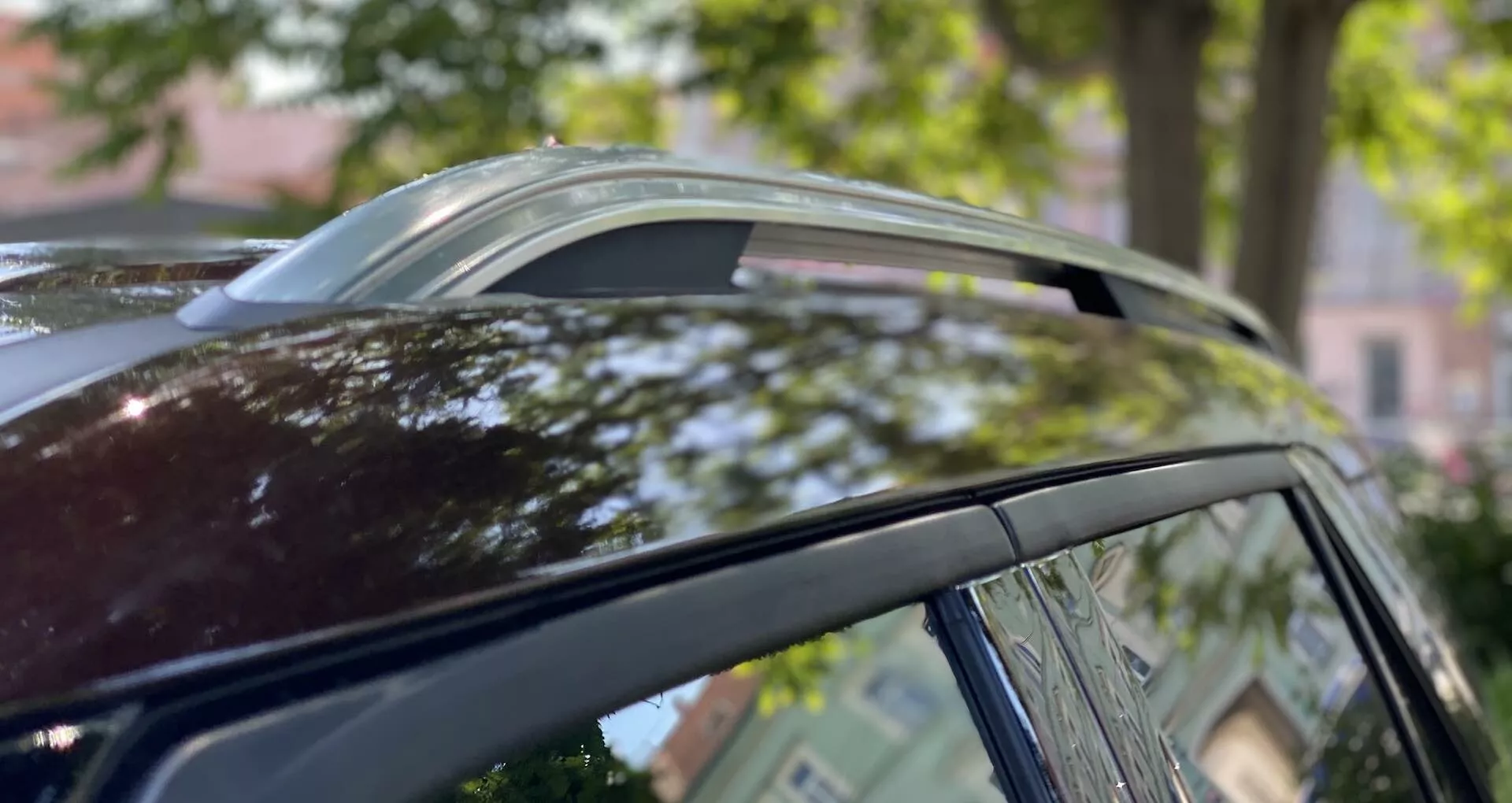 Pogled od blizu na streho Ford Explorer 2019 iz nizkega zornega kota, ki prikazuje panoramsko strešno okno v prezračevanem položaju, kovinske obrobe oken in strešne nosilce proti zamegljenemu ozadju dreves in arhitekture.