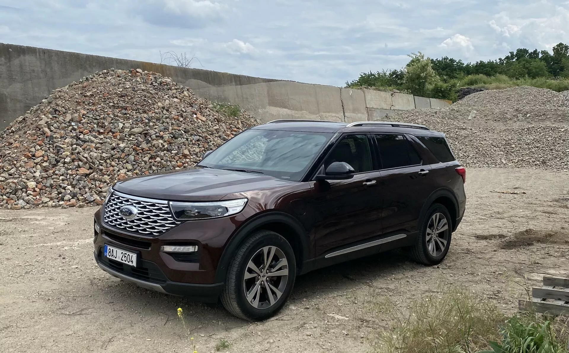 Slika prikazuje Ford Explorer 2019, sodoben SUV, parkiran na neravnem terenu, ki spominja na gradbišče ali območje za vožnjo po brezpotjih. Posneta iz treh četrtin perspektive, fotografija prikazuje sprednji in bočni del vozila, vključno z rešetko, žarometi, sprednjim odbijačem, pokrovom motorja, aluminijastimi platišči, sprednjimi in zadnjimi vrati, stranskimi ogledali z integriranimi smerniki, blatnikom in okni. Avto je obrnjen proti desni, kovinska barva pa je jasno vidna, kar omogoča celovit pogled na ta dobro vzdrževani avto z vzdolžnimi strešnimi nosilci, ki nakazujejo dodatno nosilnost.