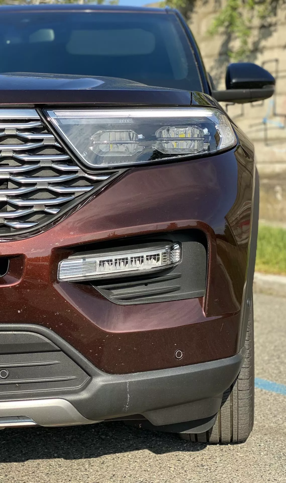 Posnetek od blizu, ki prikazuje sprednjo levo četrtino modernega bordo Ford Explorerja 2019, prikazuje zapleteno kromirano mrežo, elegantno LED sprednjo luč z dnevnimi lučmi, ohišje za meglenko s kromiranim dodatkom in kovinski zaključek, ki se sveti na soncu.