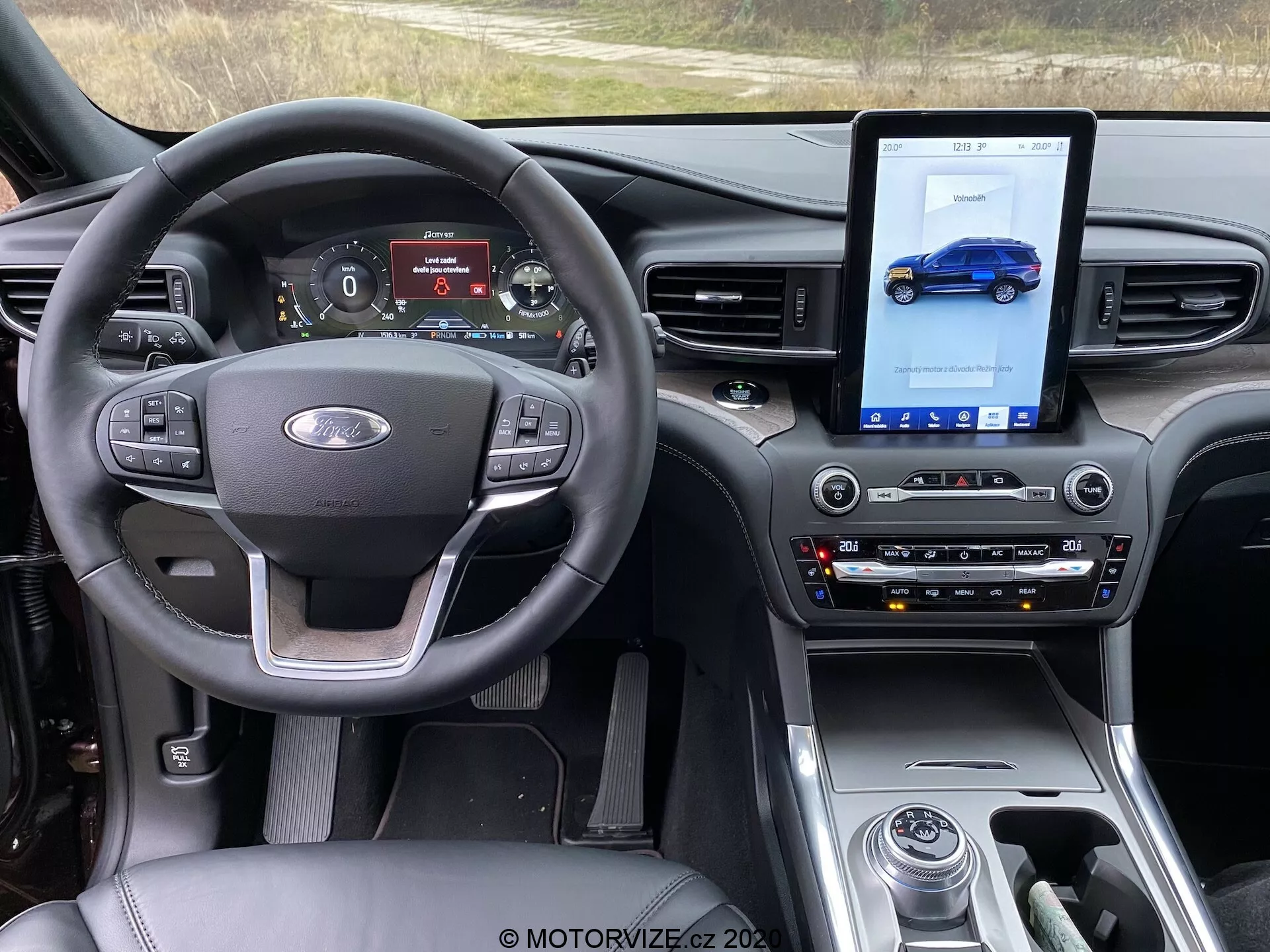 Notranjost Ford Explorerja 2019 s poudarkom na sprednji kabini iz perspektive sedeža voznika. Slika prikazuje volan z gumbi za nadzor glasnosti, glasovnimi ukazi in tempomatom, skupaj z emblemom Ford v sredini. Digitalna instrumentna plošča prikazuje merilnik hitrosti, tahometer, vozni način in stanje goriva. Sredinska konzola ima velik vertikalni zaslon na dotik za informacijsko-zabavni sistem, ki prikazuje modri avto, tradicionalne kontrole za nastavitve klime in avdio sistema ter prestavno ročico. Armaturna plošča ima sodoben dizajn s sijajnimi površinami, mat površinami in detajli iz brušenega kovine, kar povečuje sodoben, premium videz. Tehnološke funkcije vključujejo multifunkcijski volan, digitalno instrumentno ploščo, informacijsko-zabavni sistem na dotik, avtomatsko klimatsko napravo, izbirnik voznega načina in nadzor temperature sedežev.