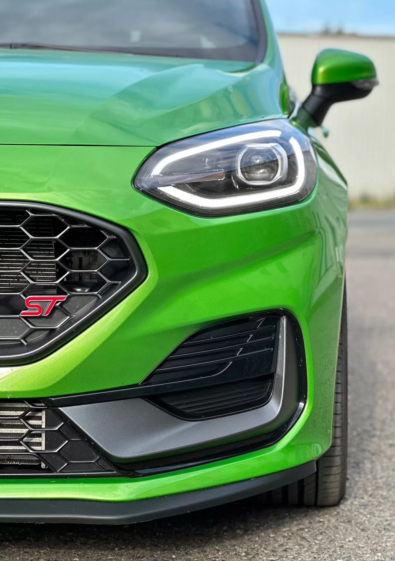 Približni pogled na sprednji levi del Ford Fieste ST 2022 v živahni zeleni, ki poudarja zmogljivostno usmerjen rob, satovje vzorčasto rešetko za optimalni pretok zraka, napreden dizajn LED žarometov, aerodinamični stranski zračnik in sprednji odbijač za boljšo usmerjanje zraka.