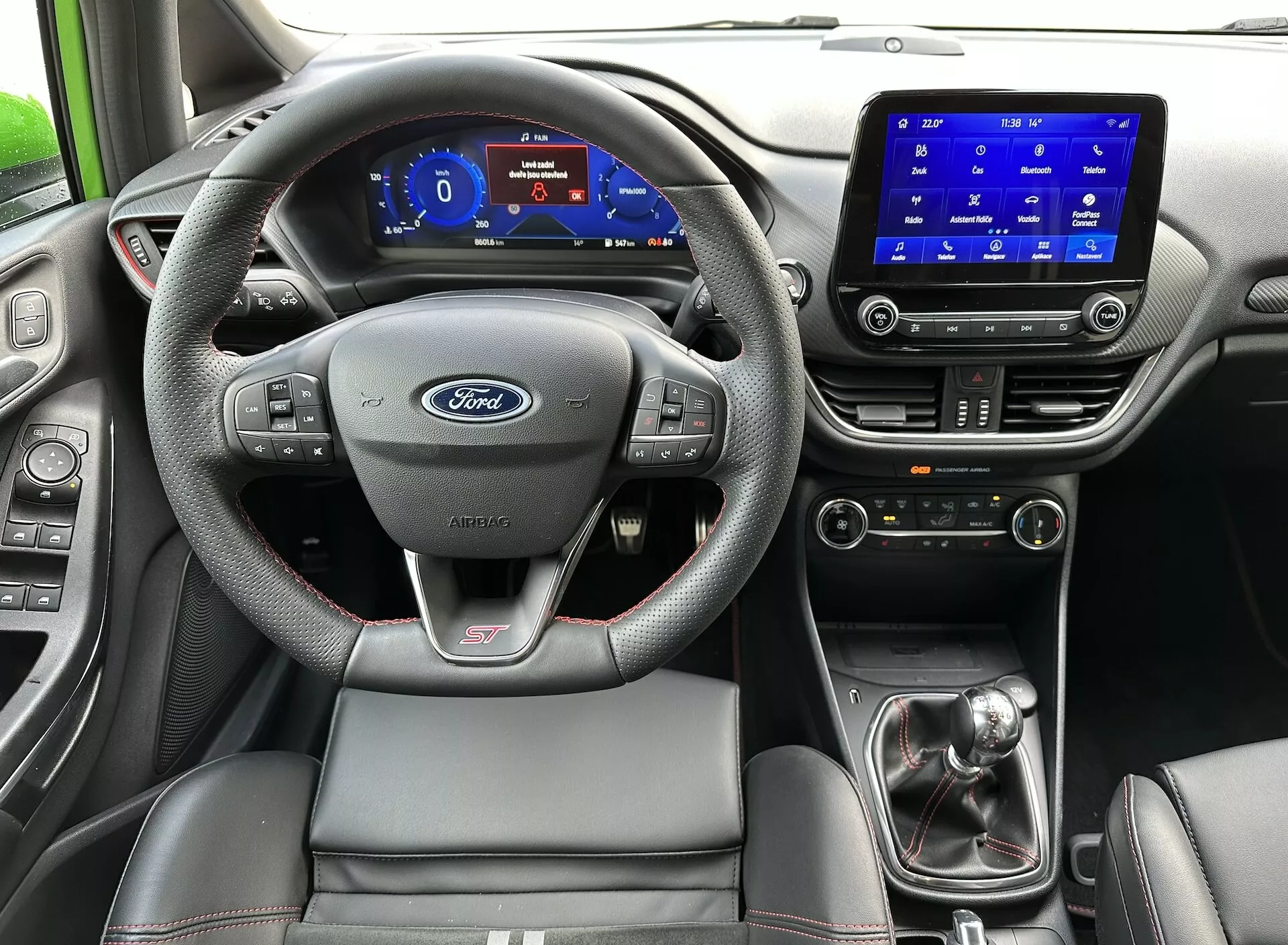 Notranjost Ford Fieste 2022 iz perspektive zadnjega sedeža, poudarja volan z logotipom Ford in značko ST, digitalni instrumentalni panel, zaslon na dotik za informacijsko-zabavni sistem, ročico za prestavljanje v ročnem načinu in športni sedež voznika s kontrastnim šivanjem.