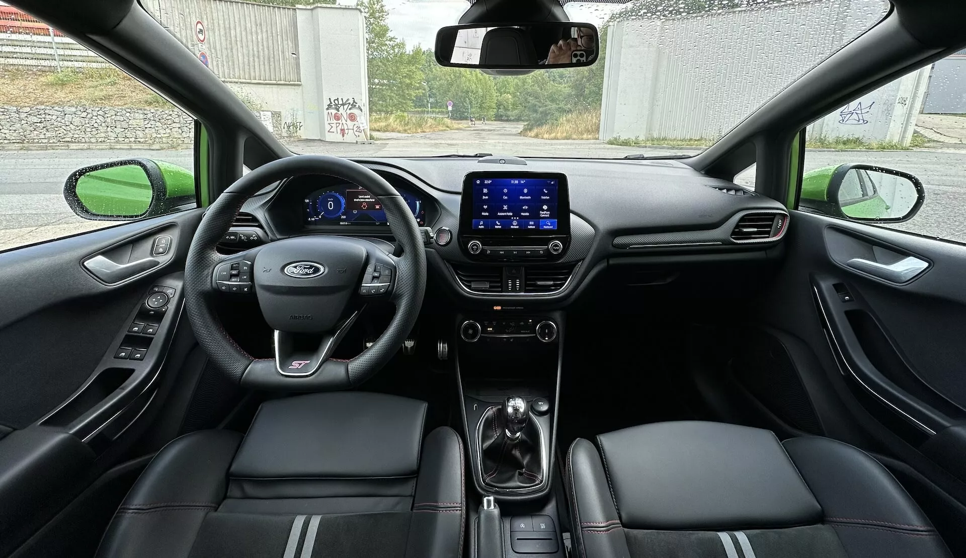 Pogled na notranjost Ford Fieste 2022 z zadnjega sedeža, ki prikazuje celoten kokpit z digitalno armaturo, večnamenskim volanom in usnjenimi sedeži z okrasnimi šivi