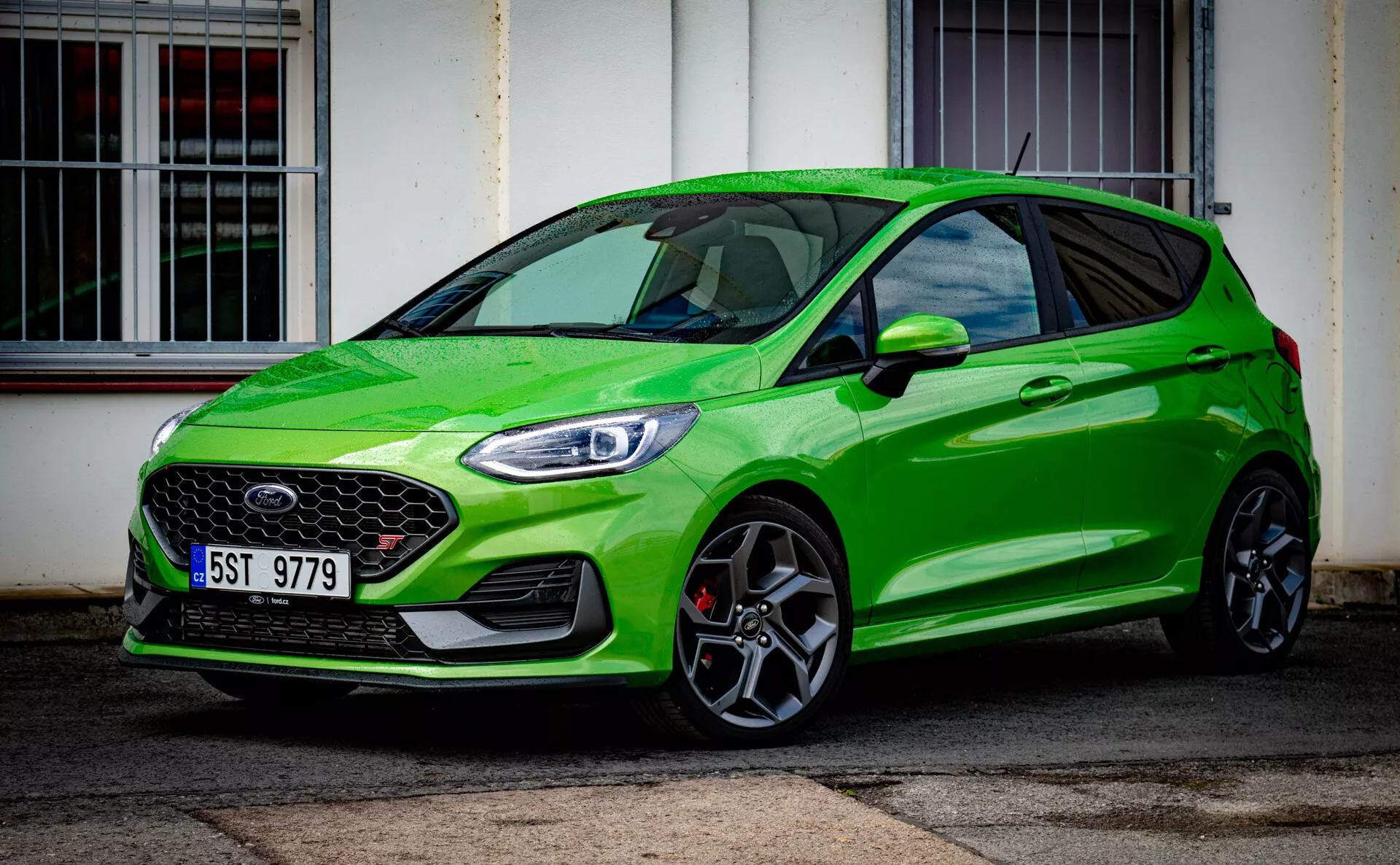 Svetlo zelen Ford Fiesta moderni model hatchback letnik 2022 poudarja športno držo s proeminentno sprednjo rešetko in elegantnimi LED žarometi, parkiran diagonalno pred zgradbo