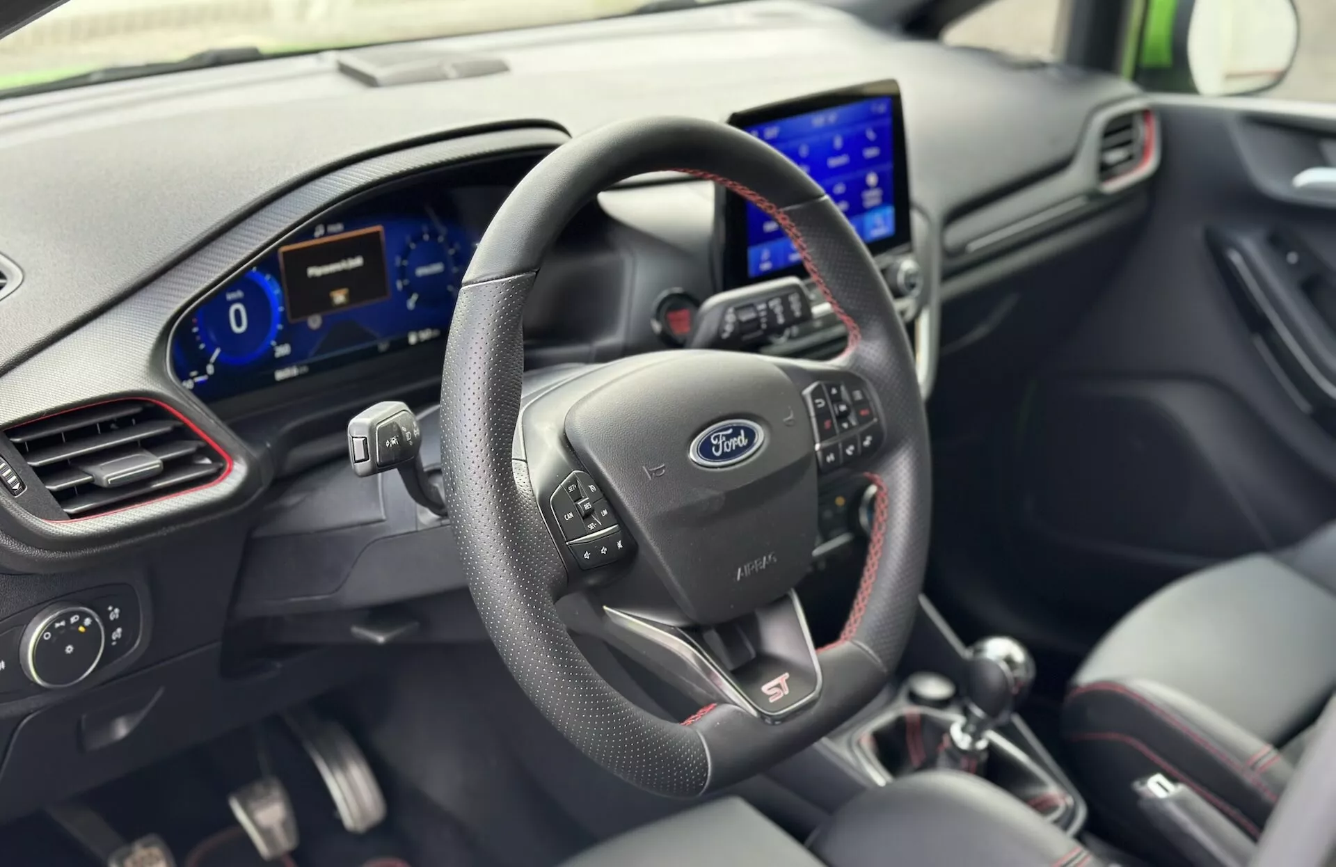 Notranjost Ford Fieste 2022 iz perspektive voznika z usnjenim volanom z nadzornimi gumbi, logotipom Ford, digitalno armaturno ploščo in sodobnim infotainment sistemom na armaturni plošči