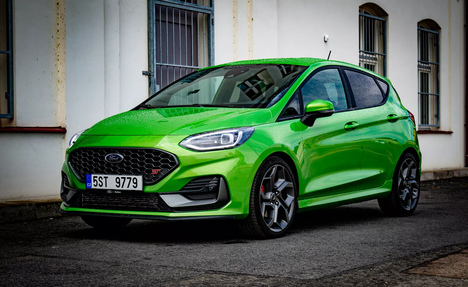 Ford Fiesta 2022 v svetlo zeleni barvi, ki prikazuje svoj športni dizajn iz nizkega sprednjega kotnega pogleda, poudarjajoč sprednji odbijač, masko, žaromete, pokrov motorja, blatnik, kolesa in odsev vetrobranskega stekla na mestni ulici po dežju