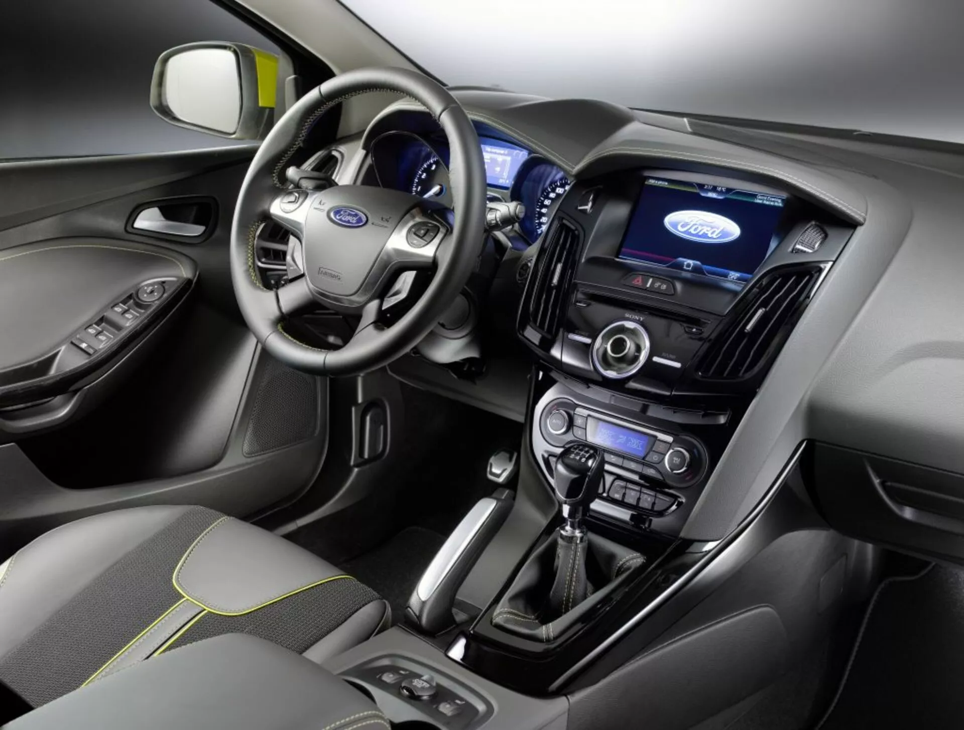 Pogled na notranjost Ford Focusa letnik 2011 z armaturno ploščo, voznikovim območjem, volanom z logotipom Ford, osvetljenim instrumentnim sklopom, centralnim zaslonom na dotik za informacijsko-zabavni sistem, klimatskimi kontrolami, avdio sistemom, avtomatskim prestavnim ročicem in sedežem sovoznika prevlečenim z blagom s šivanim vzorcem