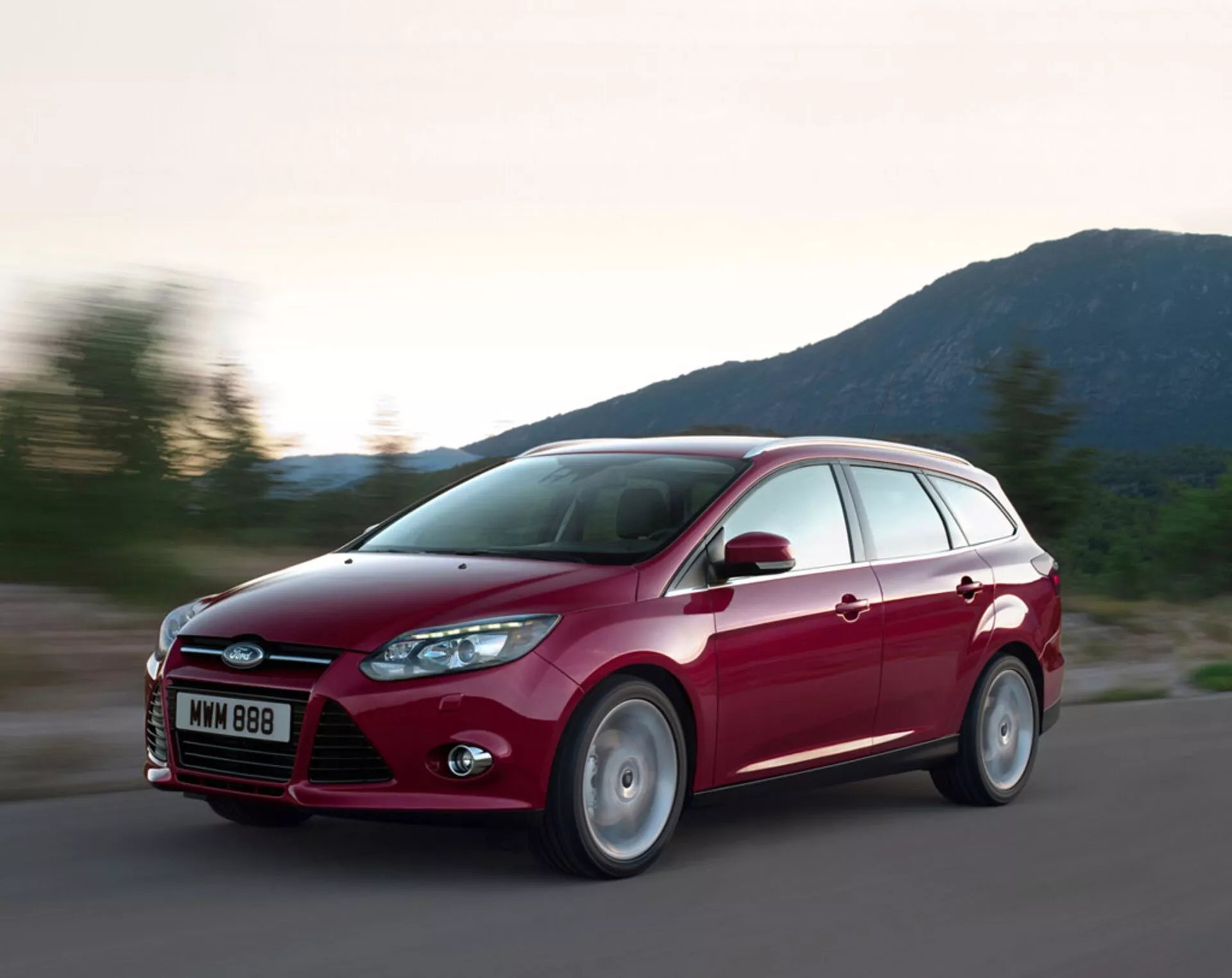 Rdeči karavanski Ford Focus letnik 2011 v gibanju po asfaltni cesti, prikazuje svoj sodoben dizajn, izrazito sprednjo rešetko z logotipom Ford, zavita prednja žarometa, oblikovane linije pokrova in lita platišča, pod večernimi svetlobnimi pogoji