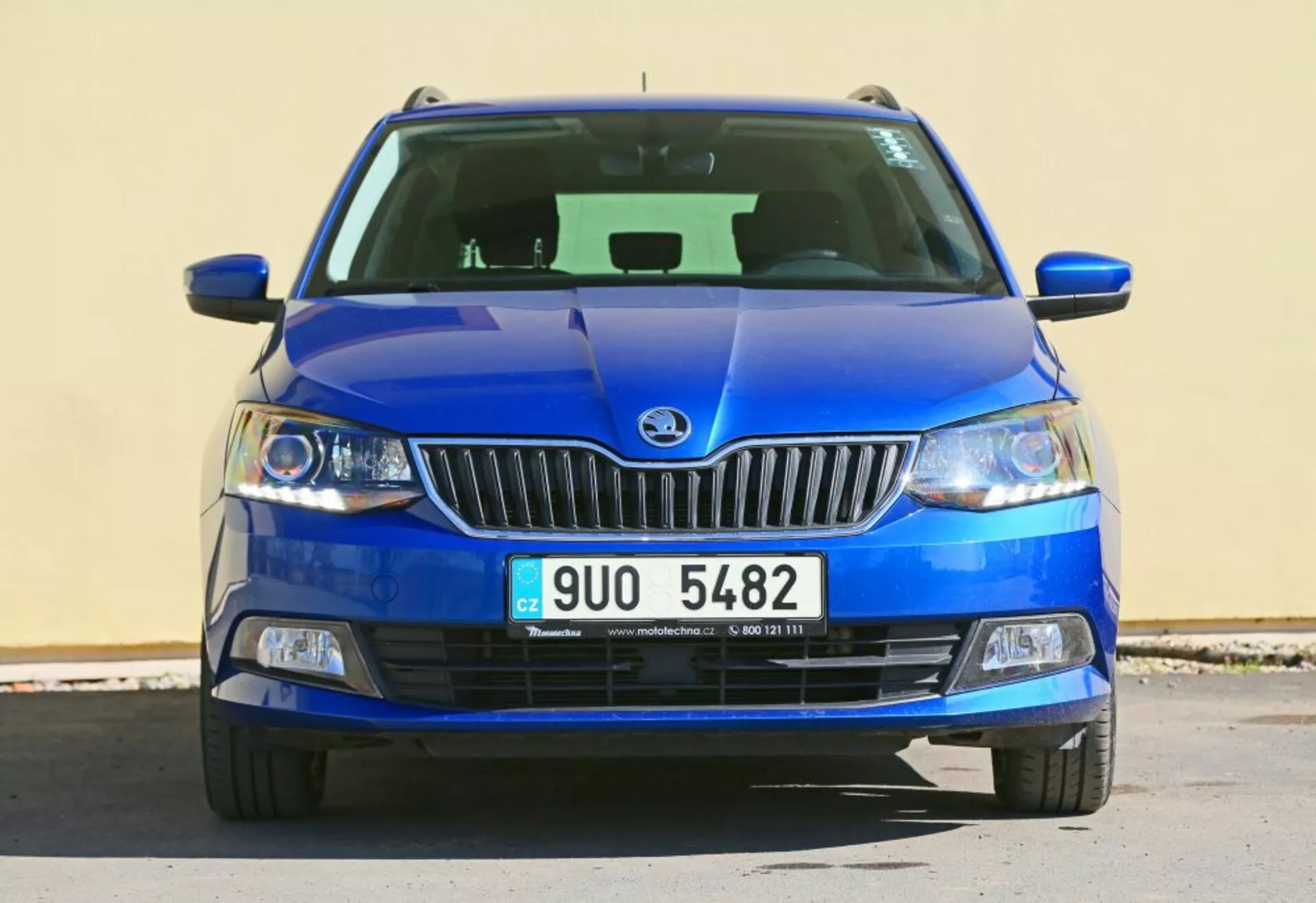 Sprednji pogled na modro vozilo Škoda z emblemo blagovne znamke na kromirani rešetki, jasnimi lečami žarometov in meglenkami v črnem ohišju, z detajlno dodelano spodnjo rešetko in kiparsko oblikovanim pokrovom motorja.