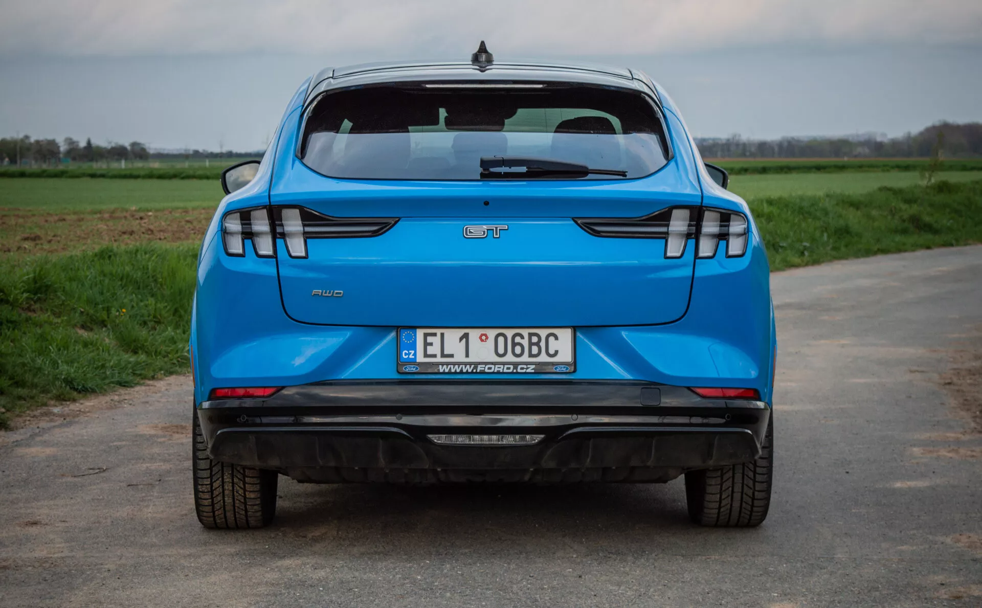 Zadnji pogled na modri Ford Mustang-Mach-E letnik 2020 na asfaltni cesti z zelenimi polji v ozadju, ki prikazuje ključne značilnosti, kot so logotip proizvajalca, ime modela, značka stopnje opreme, poljska registrska tablica, dvojni izpušni sistem, sodobne LED zadnje luči, senzorje za parkiranje in visoko nameščeno stop luč.
