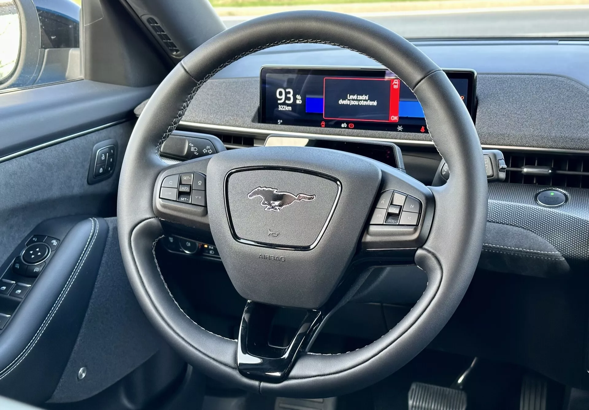 Notranjost Ford Mustang Mach-E 2020, ki poudarja sodoben volan s tremi kraki z multifunkcijskimi gumbi in digitalno instrumentno ploščo iz perspektive voznika, prikazuje kontrole, emblem in namige navigacije.