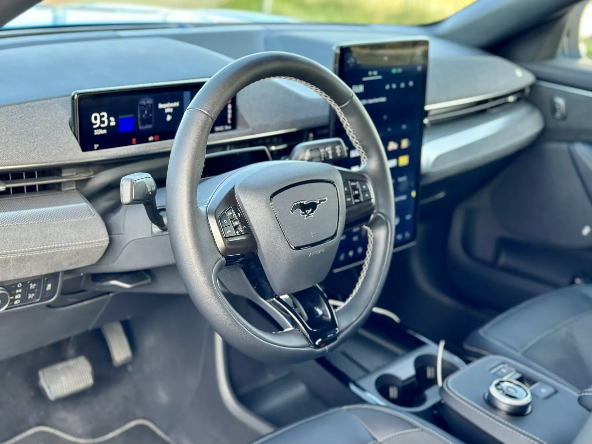 Notranji pogled na Ford Mustang Mach-E 2020 s fokusom na voznikovo kabino iz perspektive sprednjega sopotnika, predstavlja volan z emblemo Mustang, digitalno armaturno ploščo v načinu parkiranja z dometom 93 milj, in vertikalno orientiran centralni zaslon na dotik, integriran z naprednimi zmogljivostmi informacijsko-zabavnega sistema.