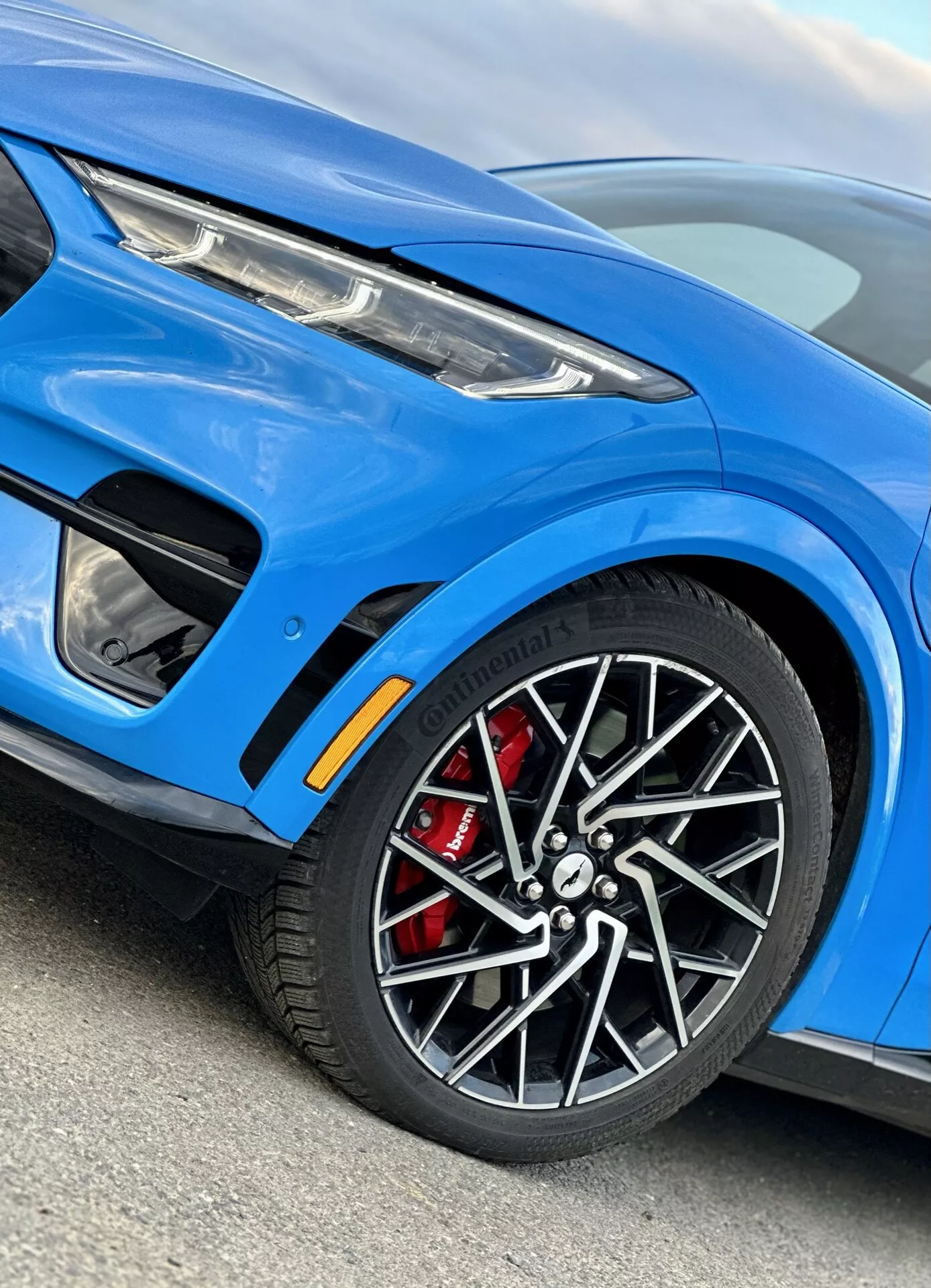 Podroben posnetek iz bližine Ford Mustang Mach-E 2020, ki prikazuje levo sprednjo stran s poudarkom na zlitinskem kolesu z rdečo zavorno čeljustjo, elegantnim sprednjim blatnikom z ventilacijsko režo, delni pogled na sodobni LED žaromet, športno modro barvo karoserije in dopolnitve zunanje opreme