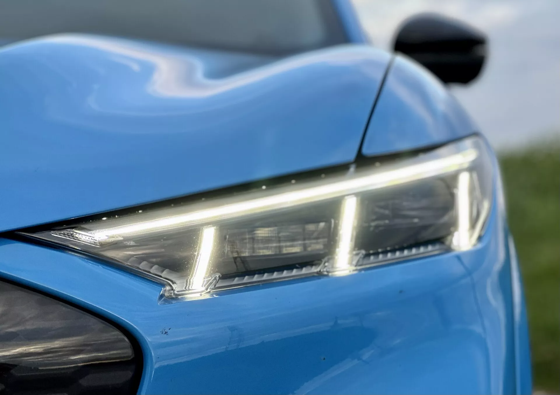 Pogled od blizu na sprednji levi del Ford Mustang-Mach-E 2020, z poudarkom na elegantni LED žaromet, živahno modro barvo, dinamične linije karoserije in del sprednjega odbijača in stranskega ogledala, kar poudarja sodobno oblikovanje in estetiko.