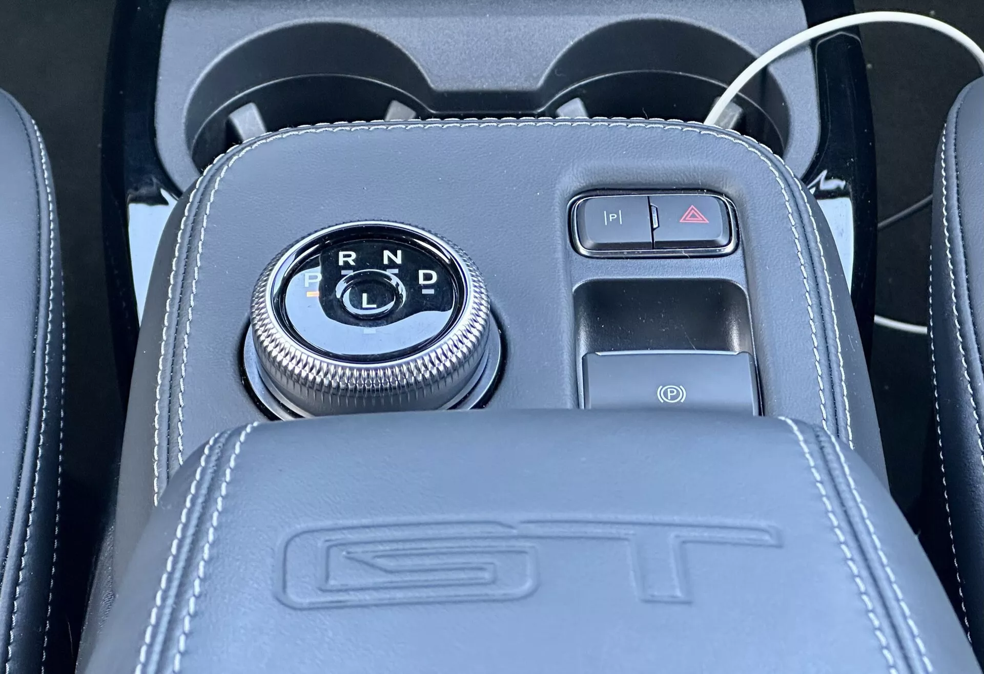 Pogled od blizu na sredinsko konzolo Ford Mustang Mach-E 2020 z rotacijskim izbirnikom prestav na sijočem črnem panelu z indikatorji prestav R, N, D, obdan z kromiranim zaključkom, zraven gumb za elektronsko zavoro parkiranja z usnjeno oblazinjenostjo, ST vtiskom, in vključujoč USB vhod in 12V vtičnico za dodatke