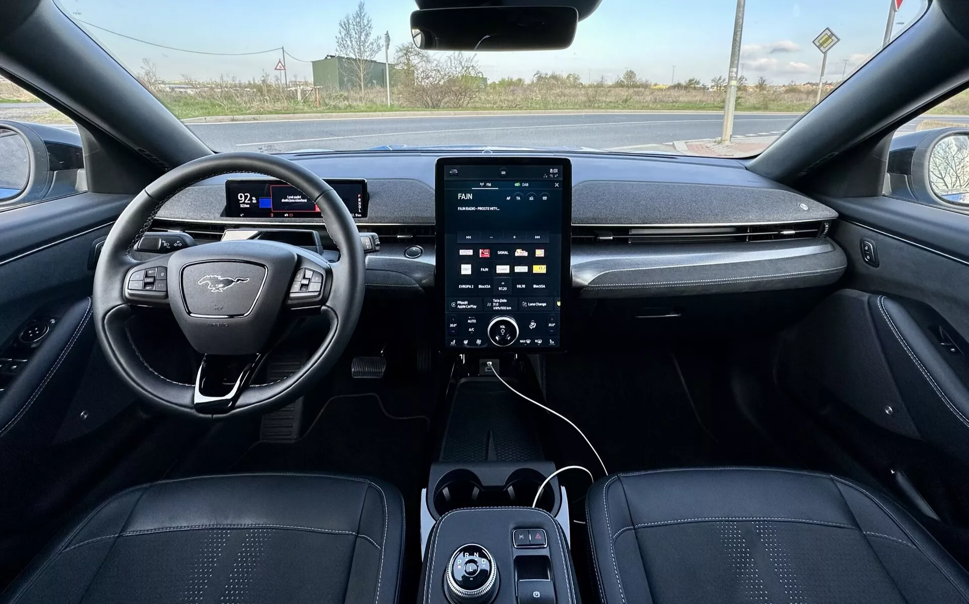 Pogled iz prve osebe notranjosti Ford Mustang Mach-E 2020, prikazuje volan na levi strani z ravnim dnom in emblemo, digitalni instrumentni sklop, velik navpičen zaslon na dotik za infotainment na sredinski konzoli z gumbi za klimatsko kontrolo spodaj, USB vhodi in razkošno temno oblazinjenje z izpostavljenimi šivi, nakazuje napredne varnostne in pomožne funkcije, z ruralnim zunanjim okoljem vidnim skozi vetrobransko steklo.