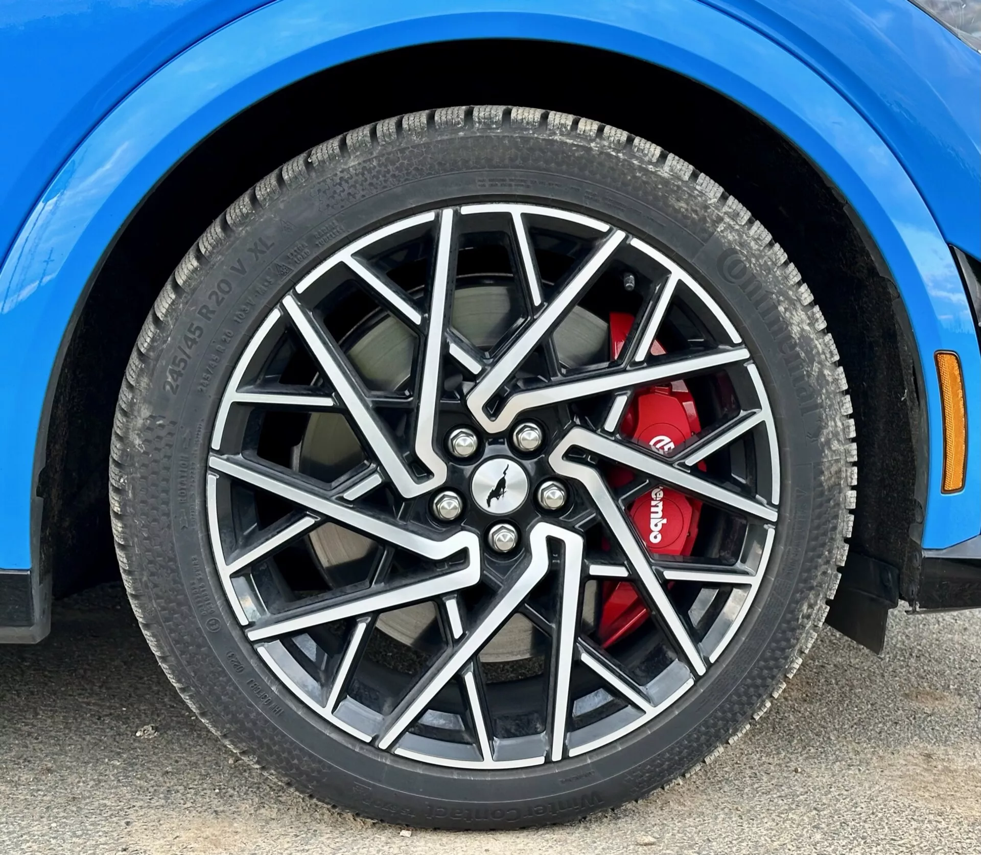 Posnetek iz blizu prednjega desnega kolesa Forda Mustang Mach-E 2020 z večkrakim zlitinskim platiščem in visokozmogljivim zavornim sistemom z rdečo zavoro, prikazuje podrobnosti pnevmatike za vse letne čase in segment drznega modrega blatnika, posneto iz nizkega kota.