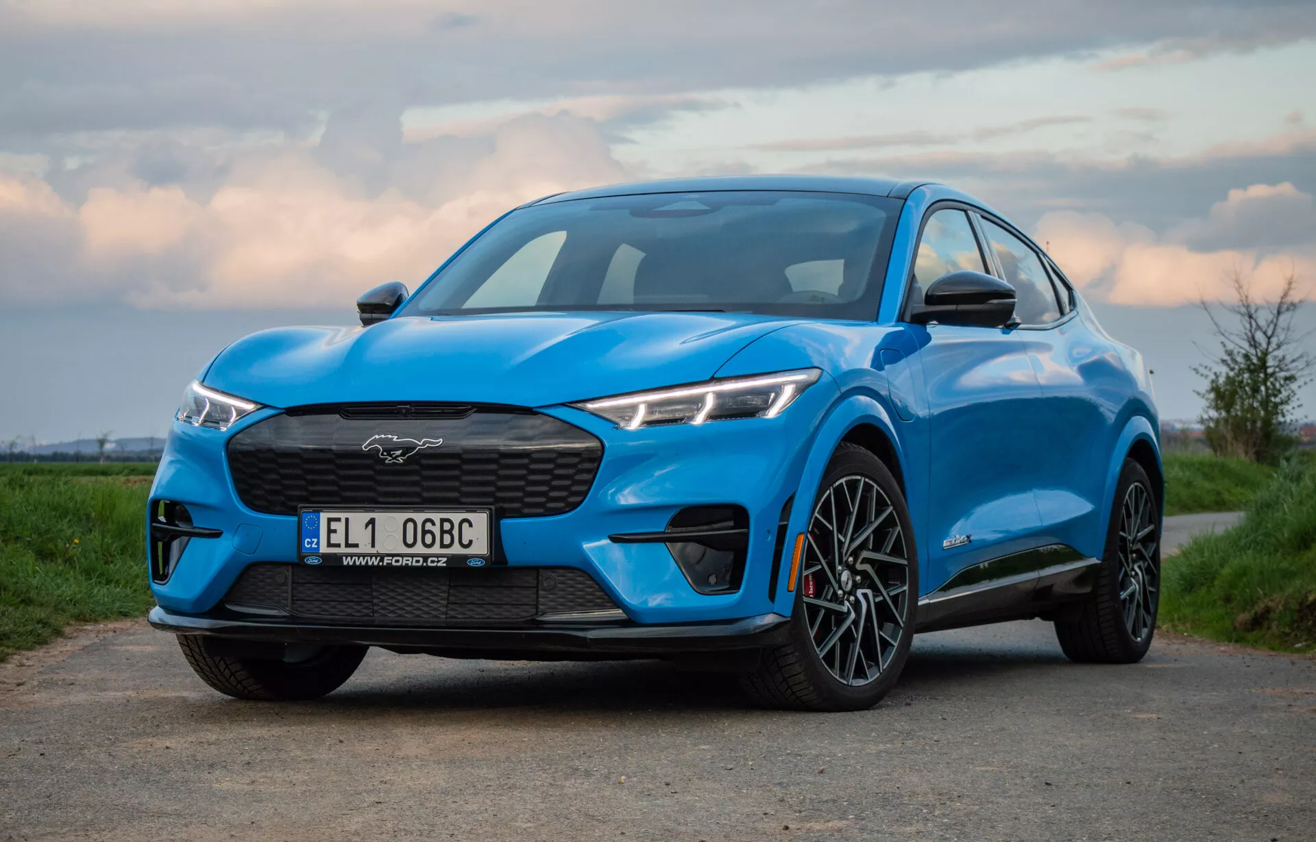 Modri Ford Mustang Mach-E 2020, fotografiran iz nizkega in rahlo stranskega kota, prikazuje rešetko, žaromete, pokrov motorja in profil električnega SUV-ja, vključno s prednjim kolesom in blatnikom, poudarjajoč njegovo zmogljivost in aerodinamično zasnovo ob zori ali mraku.