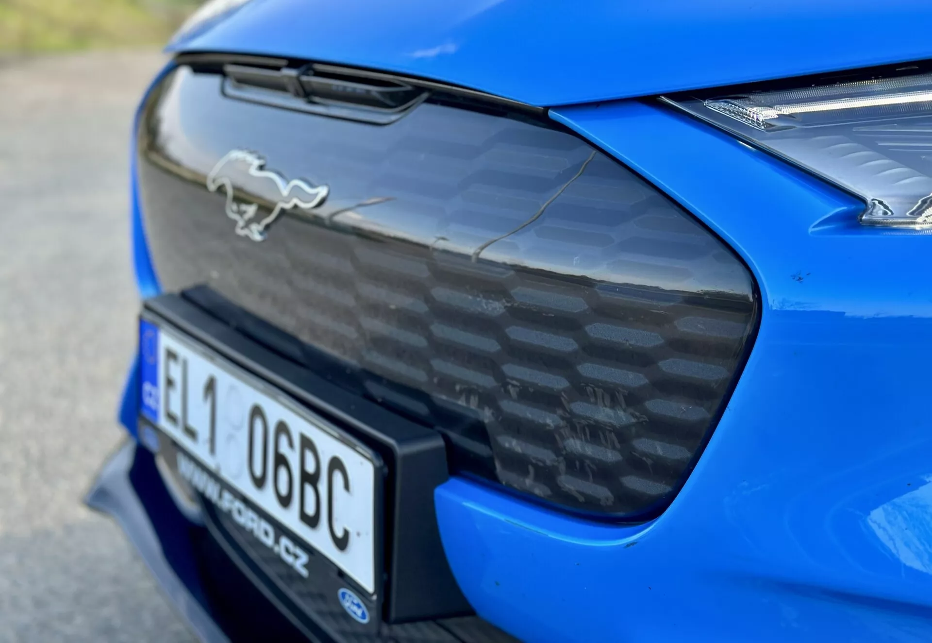 Podroben posnetek zadnjega dela modrega športnega avtomobila Ford Mustang Mach-E 2020, ki prikazuje kromiran emblem konja, spojler iz ogljikovih vlaken na zadnjem pokrovu, integrirani prostor za registrsko tablico in delni pogled na LED zadnje luči, poudarjajoč aerodinamične in zmogljivostne značilnosti avtomobila.