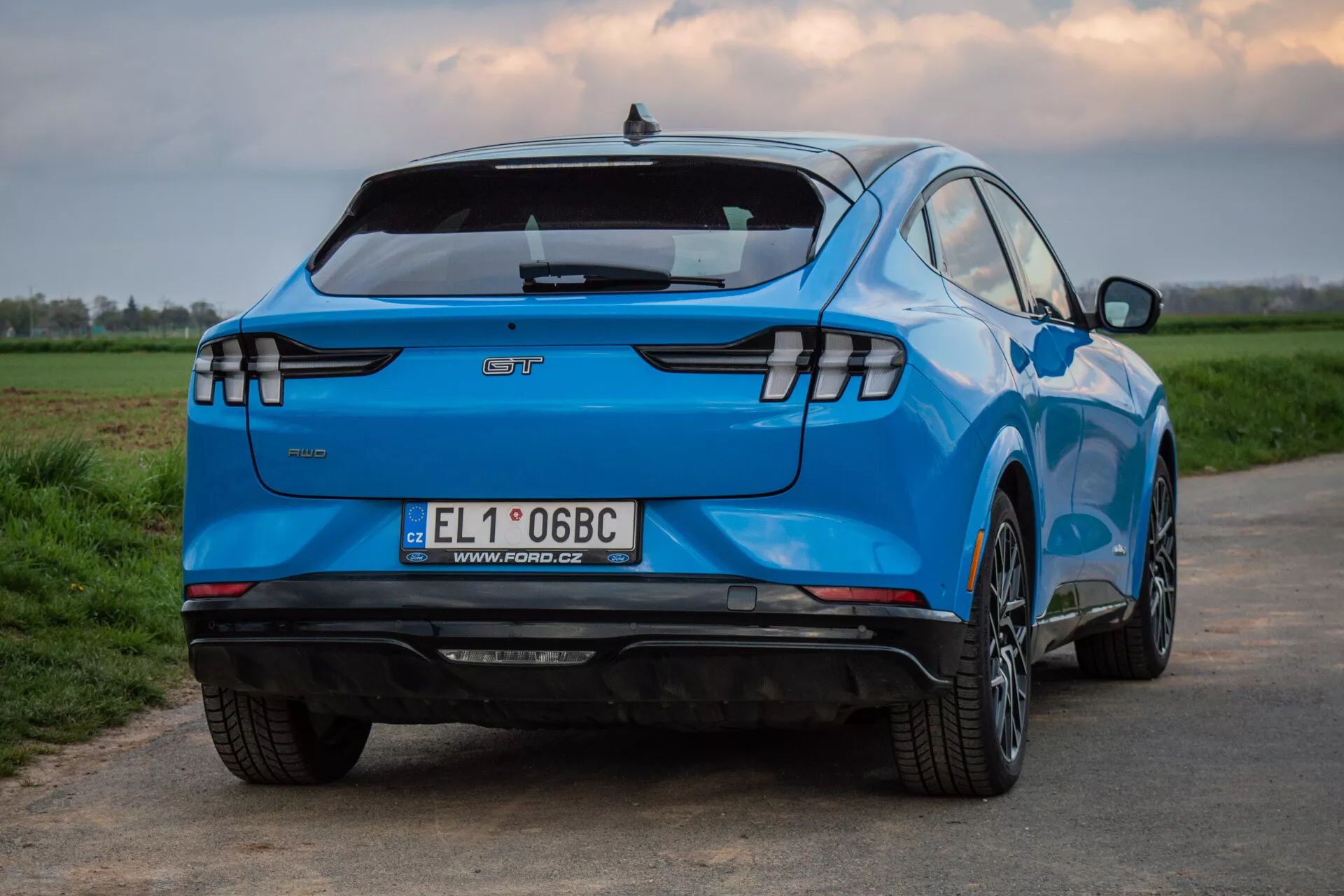 Ford Mustang-Mach-E 2020 fotografiran z zadnjega trehčetrtinskega kota, ki prikazuje njegovo značilno obliko, vključno s horizontalnimi LED zadnjimi lučmi, emblemo proizvajalca, integriranim zadnjim spojlerjem, zadnjim odbijačem opremljenim s senzorji brez vidnega izpuha, kar nakazuje status električnega vozila, evropsko registracijsko tablico, večkraka lita kolesa, živo modro barvo in aerodinamično oblikovano karoserijo, na podeželskem ozadju.