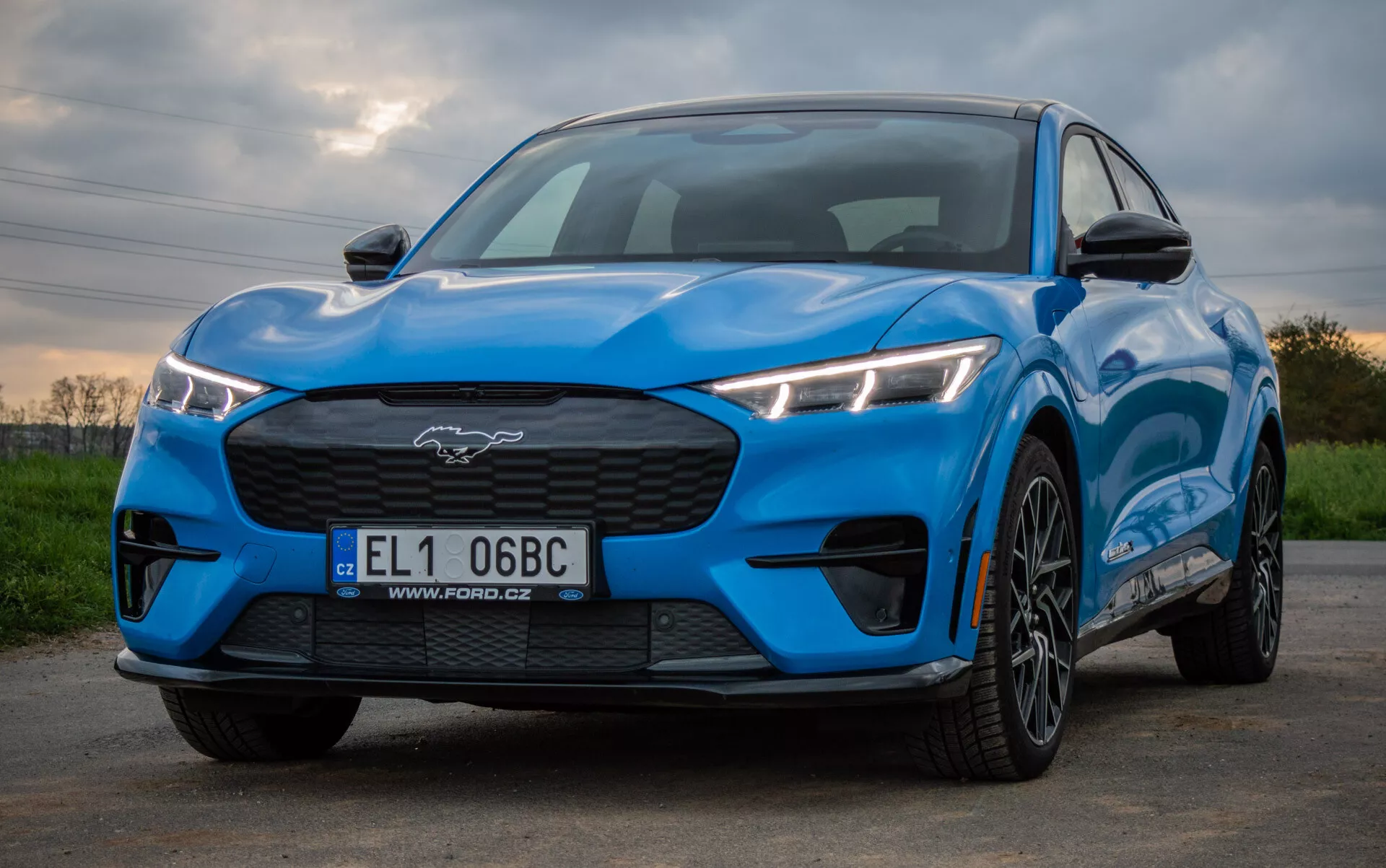 Ford Mustang Mach-E 2020 v svetlo modri barvi, ki izpostavlja športno in aerodinamično zasnovo iz nizkega sprednjega kota, poudarjajoč kotne LED žaromete, zaprto rešetko, oblikovano pokrov motorja in večkrake zlitine koles proti naravnemu ozadju.