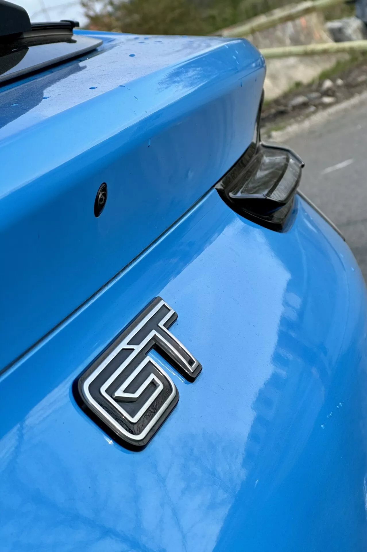 Podroben posnetek zadnjega dela modrega Ford Mustang Mach-E letnik 2020 s poudarkom na kovinski znački GT, ukrivljenem prtljažniku in aerodinamičnem zadnjem spojlerju s črnim zaključnim pokrovčkom proti nežno zamegljenem ozadju.