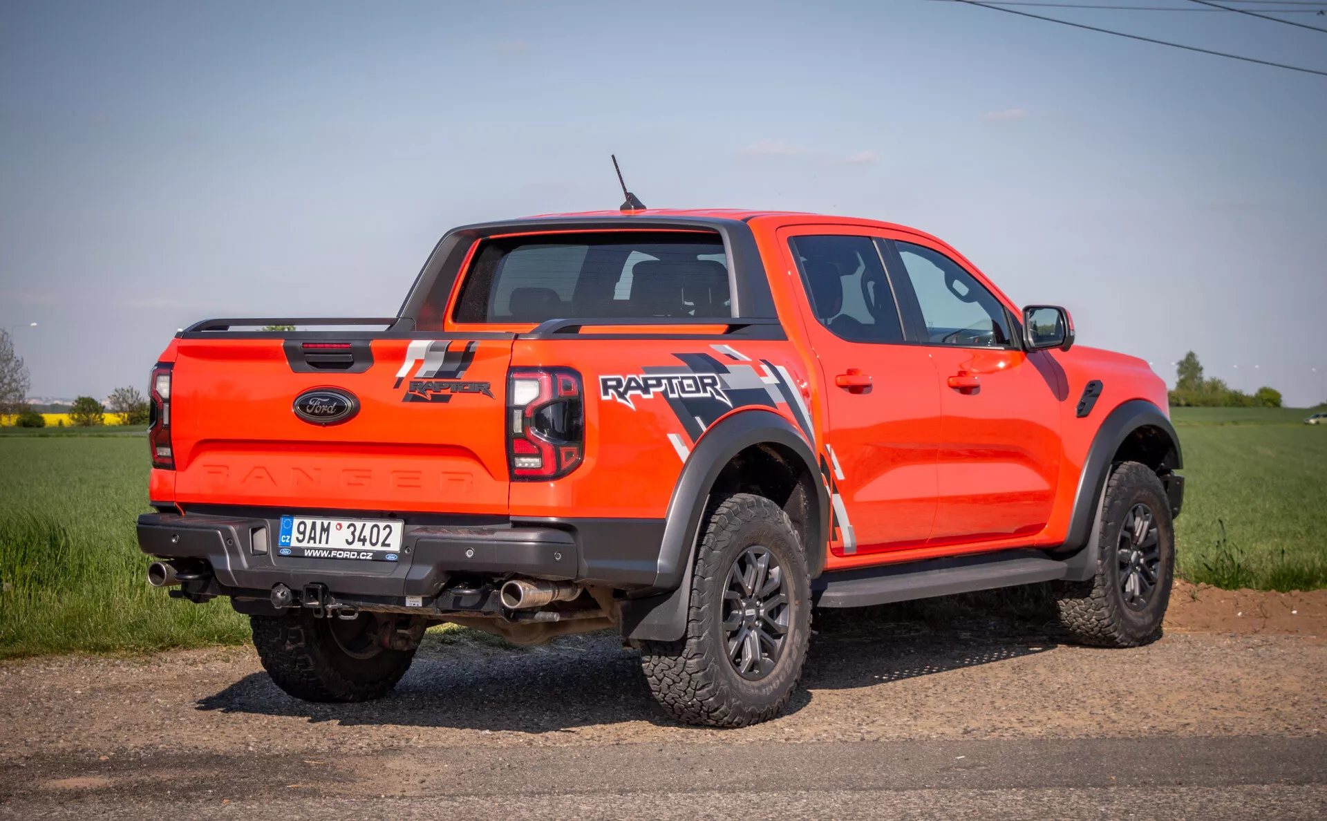 Svetlo oranžno pickup vozilo Ford Raptor 2022 iz stransko-zadnjega zornega kota, na prostem na polju, ki prikazuje svoje sposobnosti za terensko vožnjo z visoko oddaljenostjo od tal, kabino s štirimi vrati, velike črne obrobe kolesnih lokov, črna platišča z več kraki in grobe terenske pnevmatike, ter zadaj blagovno znamko 'RAPTOR' na prtljažnih vratih.
