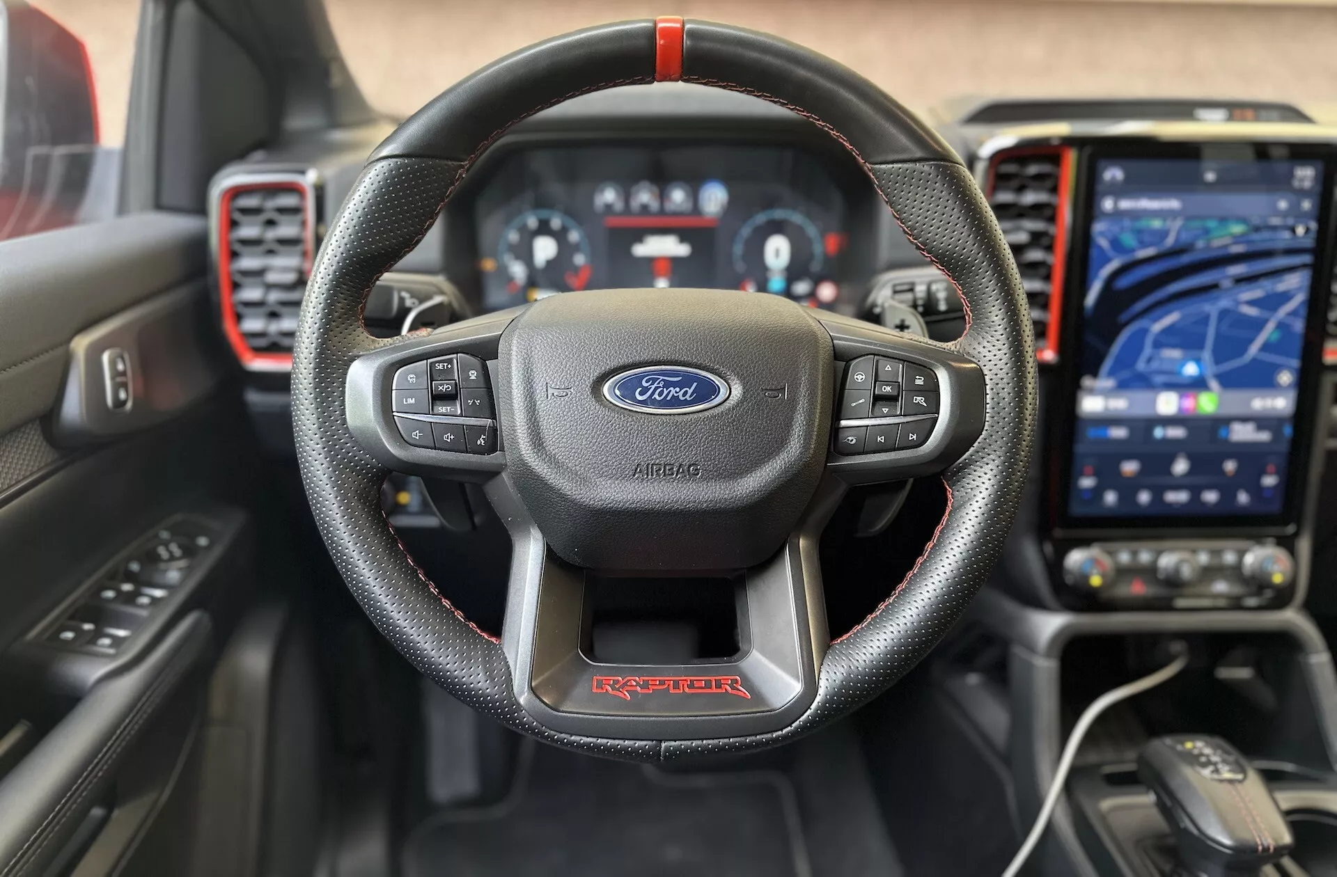 Notranji sprednji pogled na Ford Ranger Raptor 2022, ki poudarja usnjeno, trokrako volansko kolo z rdečimi šivi in Fordovim logotipom, digitalni instrumentni zaslon, sodoben dotični informacijsko-zabavni sistem v načinu navigacije, športno armaturno ploščo z rdečimi poudarki in napredne pripomočke za voznika za kakovostno, tehnološko osredotočeno izkušnjo vožnje