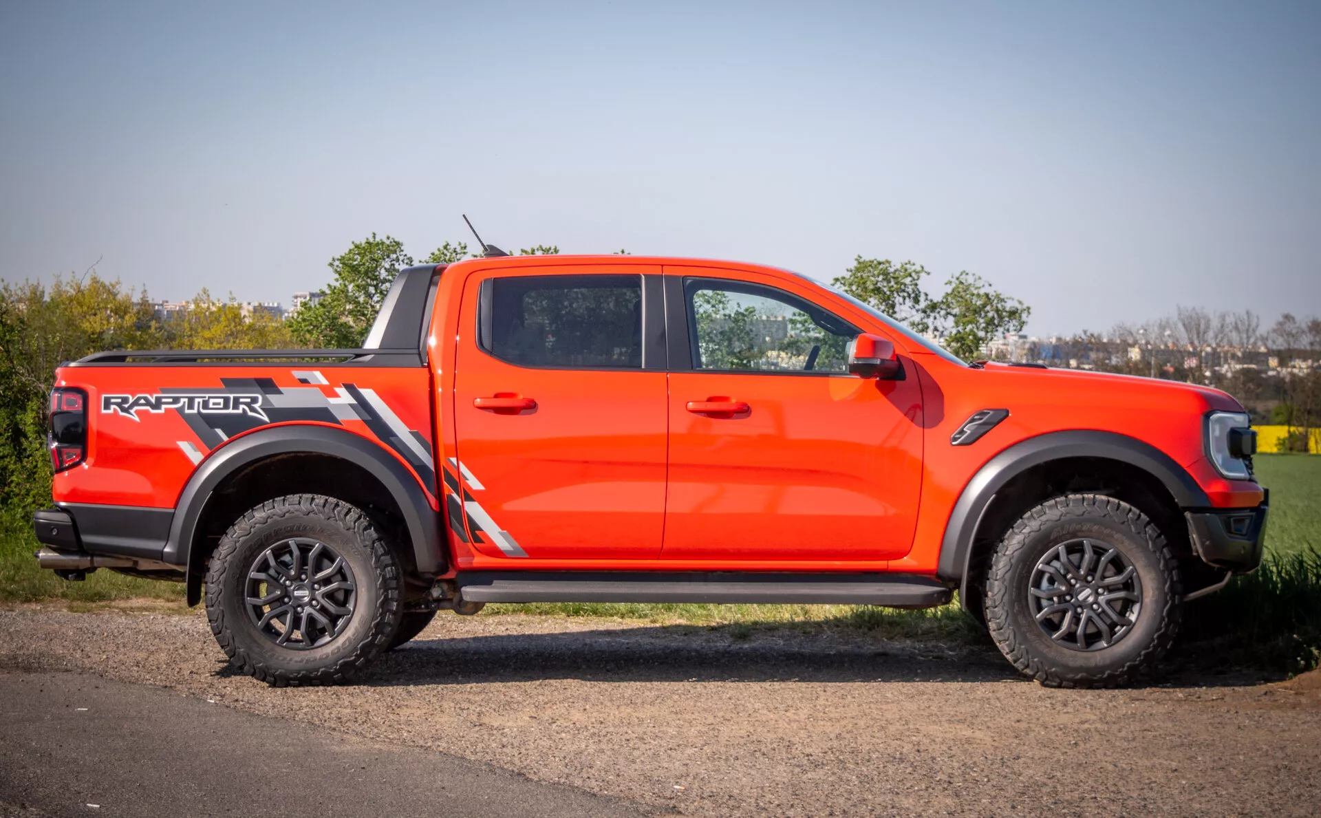 Ford Ranger Raptor letnika 2022 v živo oranžni barvi, ki kaže agresivni slog, zmogljivosti terenske vožnje z velikimi črnimi platišči in pnevmatikami z globokim profilom, parkiran na tlakovanem površju ob travi čez dan