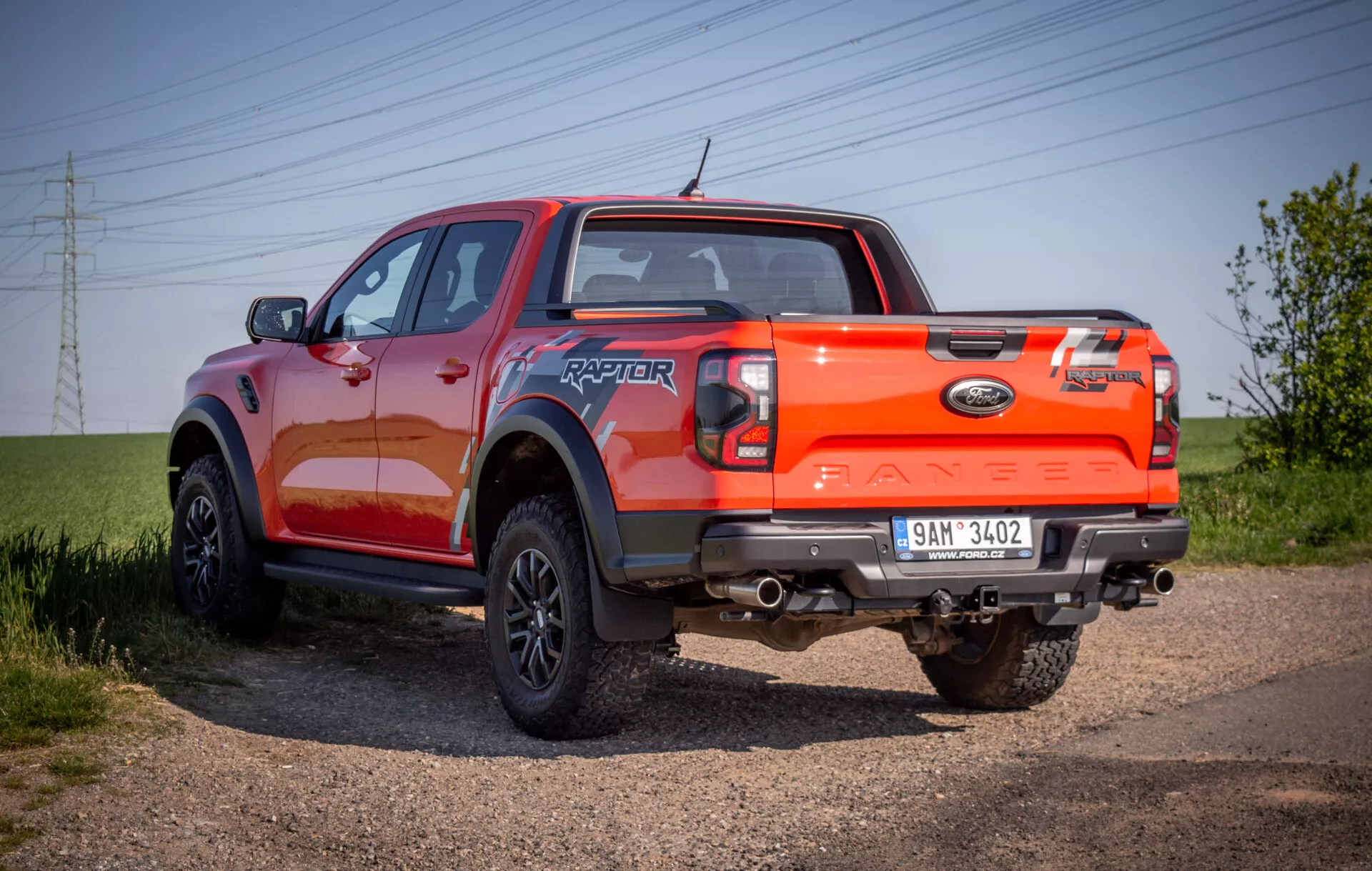 Pickup Ford Ranger Raptor 2022, prikazan na makadamski poti, ki izpostavlja njegov robusten zadnji tričetrtinski kot s poudarkom na zmogljivostih za vožnjo izven cest in živahni oranžni barvi zaključka.