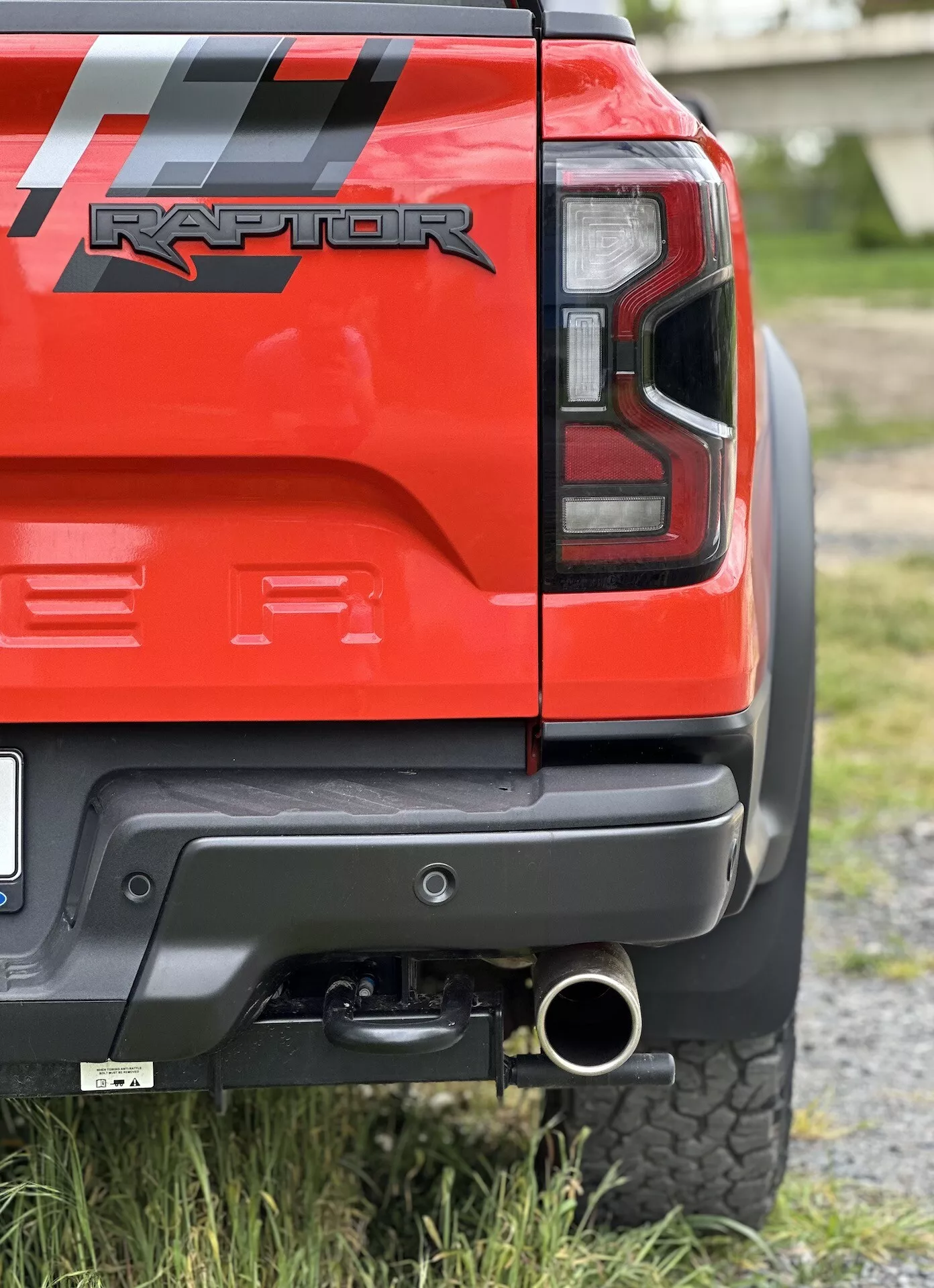 Približni posnetek zadnjega kota rdečega Ford Ranger Raptor 2022, ki prikazuje LED zadnjo luč, črni zadnji odbijač s sivim panelom, vlečno kljuko, polirano konico izpuha in pnevmatiko za vse terene, poudarja njegove sposobnosti za vožnjo izven cest in podrobnosti oblikovanja.