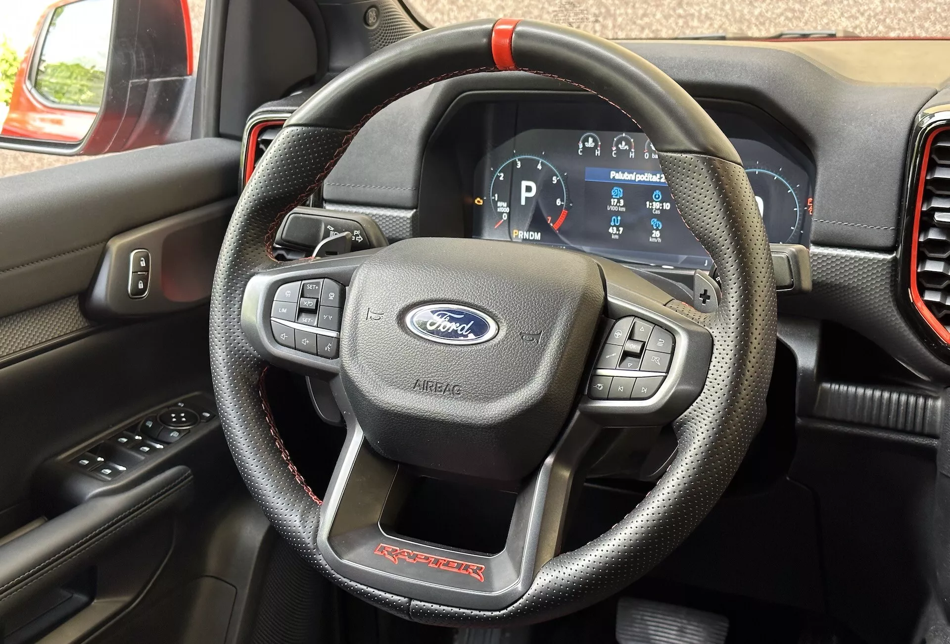 Podroben pogled na notranjost Ford Ranger Raptor 2022, osredotočen na krmilnik, ki posnema usnje z rdečimi šivi in multifunkcijskimi kontrolami, ter digitalno instrumentno ploščo, ki prikazuje vozilo v načinu parkiranja.