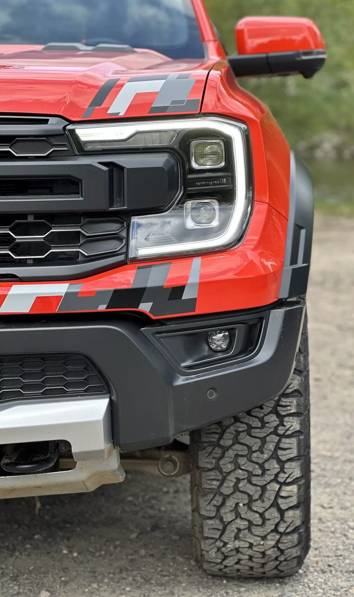Podrobn pogled na rdeči pickup Ford Ranger 2022, ki prikazuje LED žaromet, robustno rešetko, mišičast sprednji blatnik z nalepko in vzorec profila pnevmatike za vožnjo po terenu z nizkega kota, da poudari robustnost in napredno tehnologijo