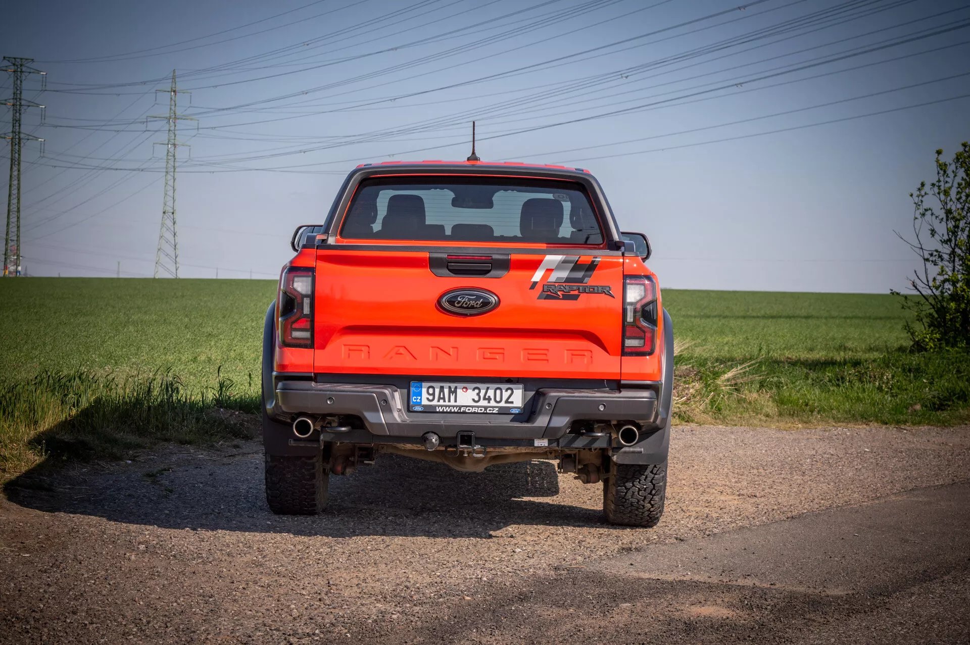 Zadnji pogled na svetlo oranžni Ford Ranger 2022 kaže celotno širino, zadnje luči, osrednje 'RANGER' blagovno znamenje in oznake posebne izdaje, s kromiranim odbijačem, ki vključuje parkirne senzorje, posnetek iz nizkega kota na makadamski cesti z ozadjem jasnega neba