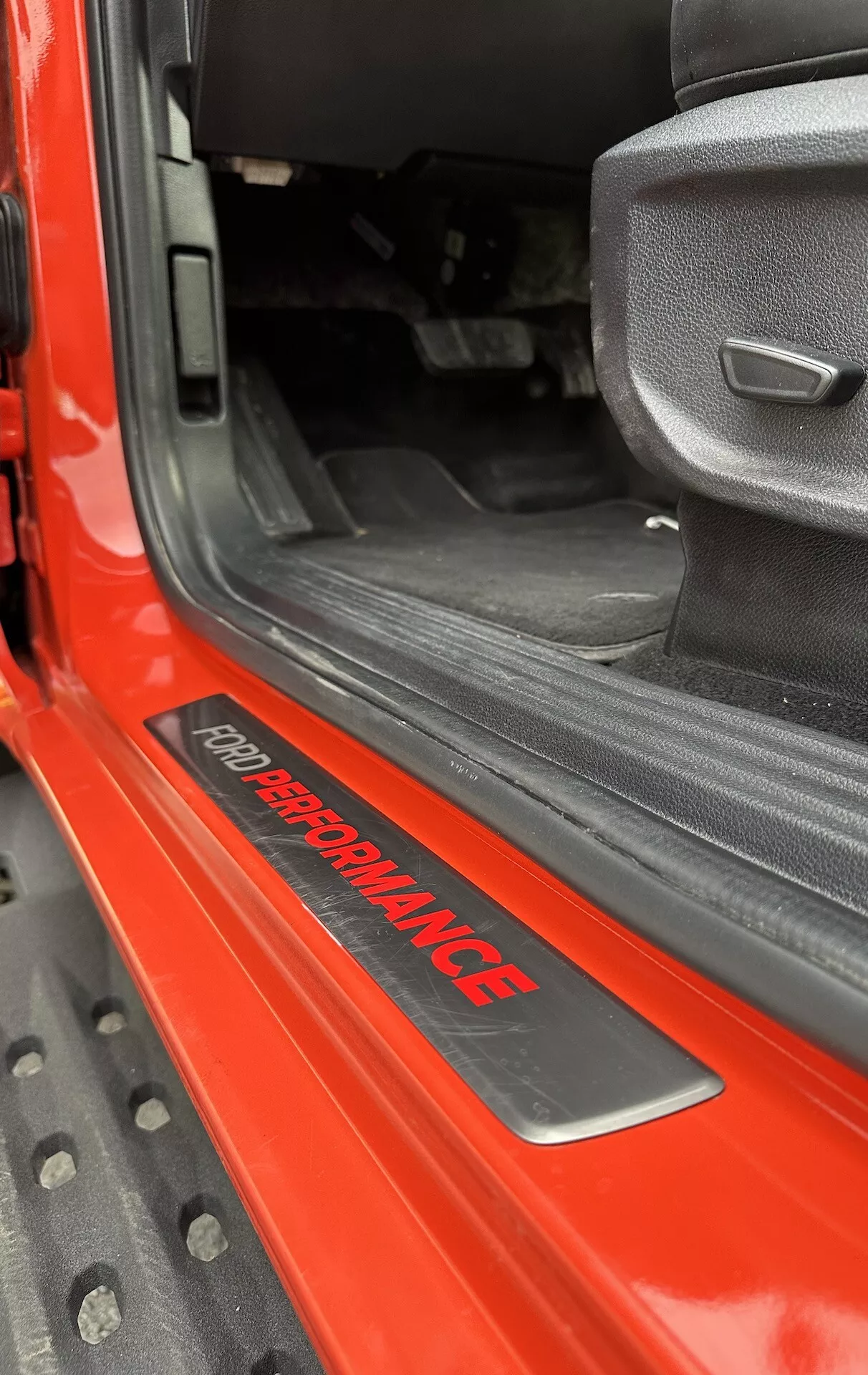 Posnetek praga vrat Ford Ranger 2022 s ploščico 'FORD PERFORMANCE', sijoč rdeč zunanji del, črna notranja preproga, sedež iz sive tkanine s športnim dizajnom in vzdržljiva črna gumijasta talna obloga