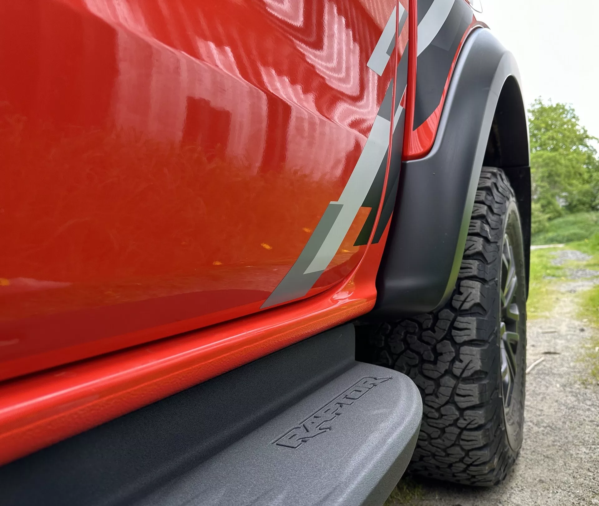 Detajlen pogled iz nizkega kota na spodnji del vrat in območje kolesa svetlo rdečega Ford Rangerja 2022, ki izpostavlja mat črni bočni korak z vgraviranim logotipom, športno nalepko na vratih in robustno pnevmatiko za terensko vožnjo.