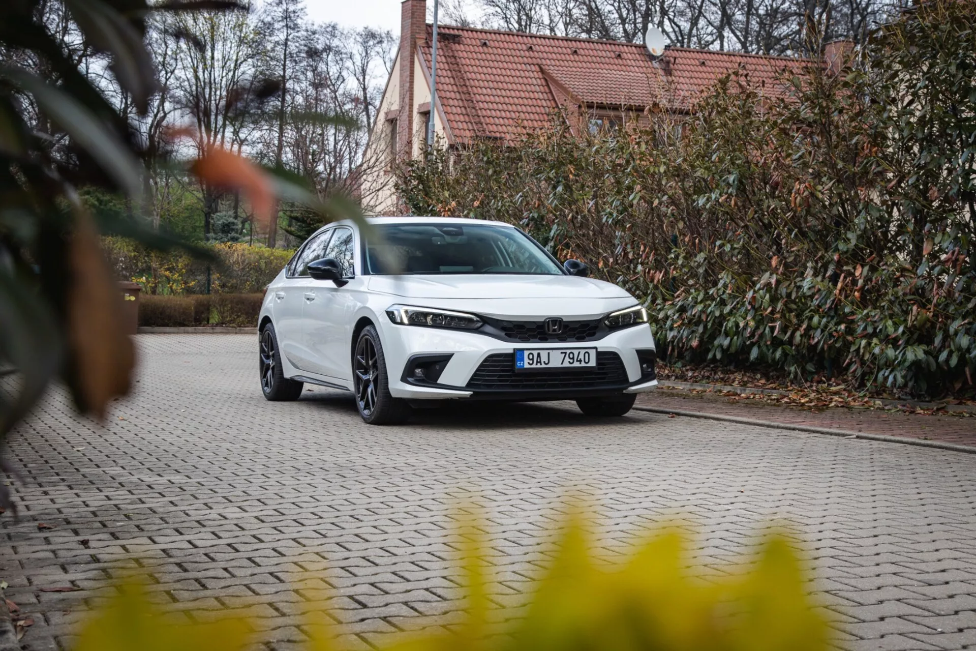 Sodobna limuzina Honda Civic 2022, parkirana na tlakovani dovozni poti z urejeno okolico in stavbo v ozadju, prikazuje sprednji žar, LED žaromete in elegantno aerodinamično obliko
