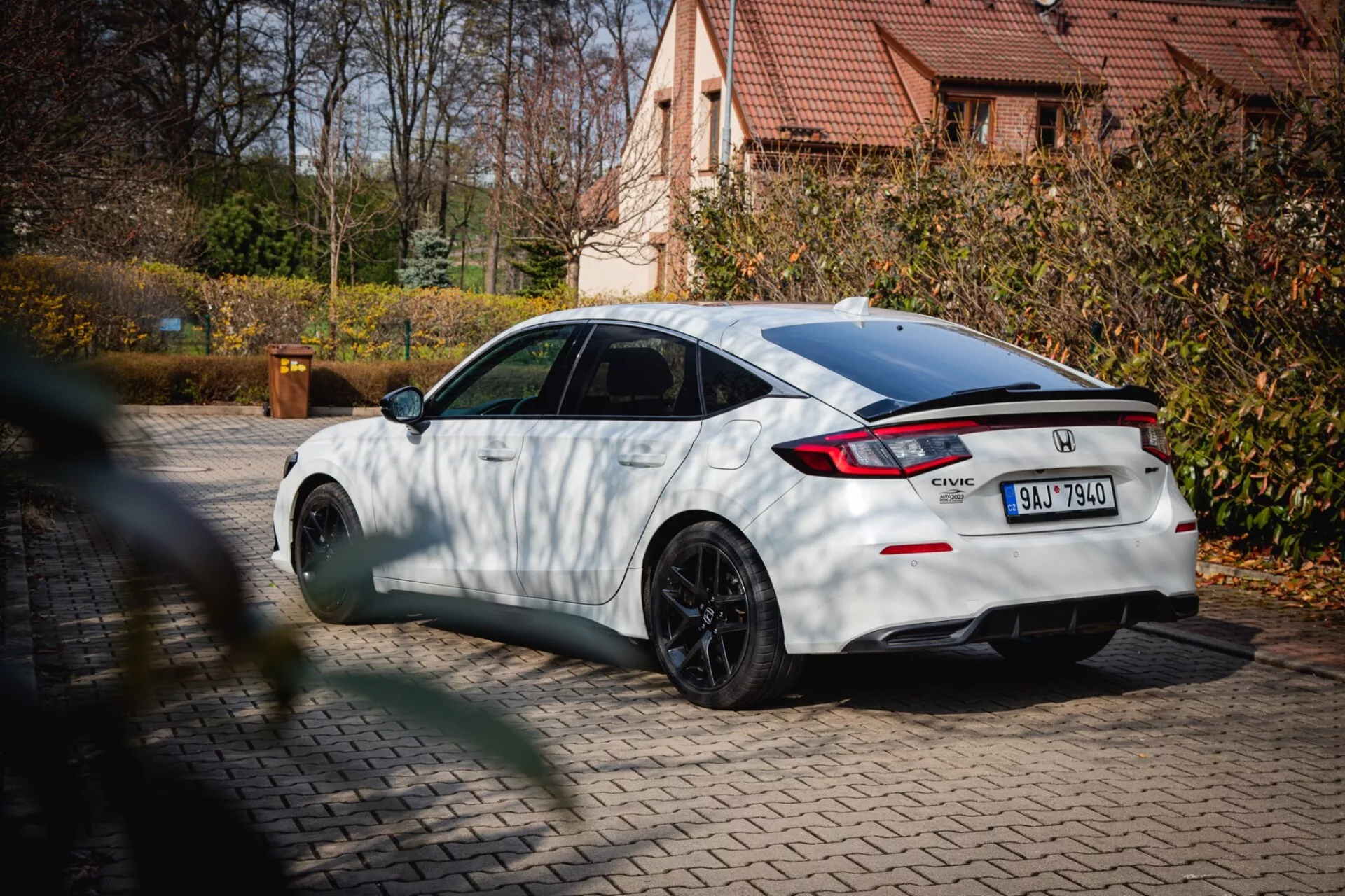 bel Honda Civic leta 2022 s črnimi večkrakimi kolesi in zadnjim spojlerjem, parkiran na tlakovcu z jesenskim listjem in stanovanjsko hišo v ozadju