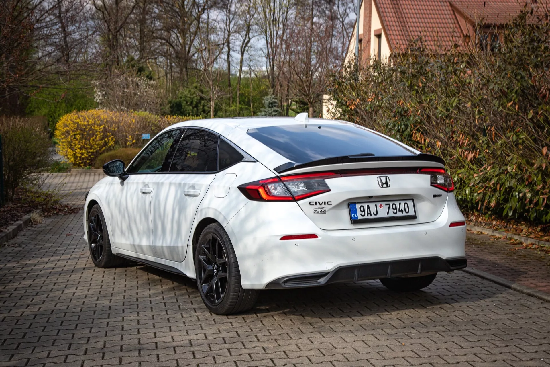 Beli Honda Civic 2022 prikazuje zadnji in delni stranski profil na opečnati dovozni poti, s padajočo streho, integriranim spojlerjem, LED zavorno lučjo, značilnimi zadnjimi lučmi in športnim odbijačem z difuzorjem
