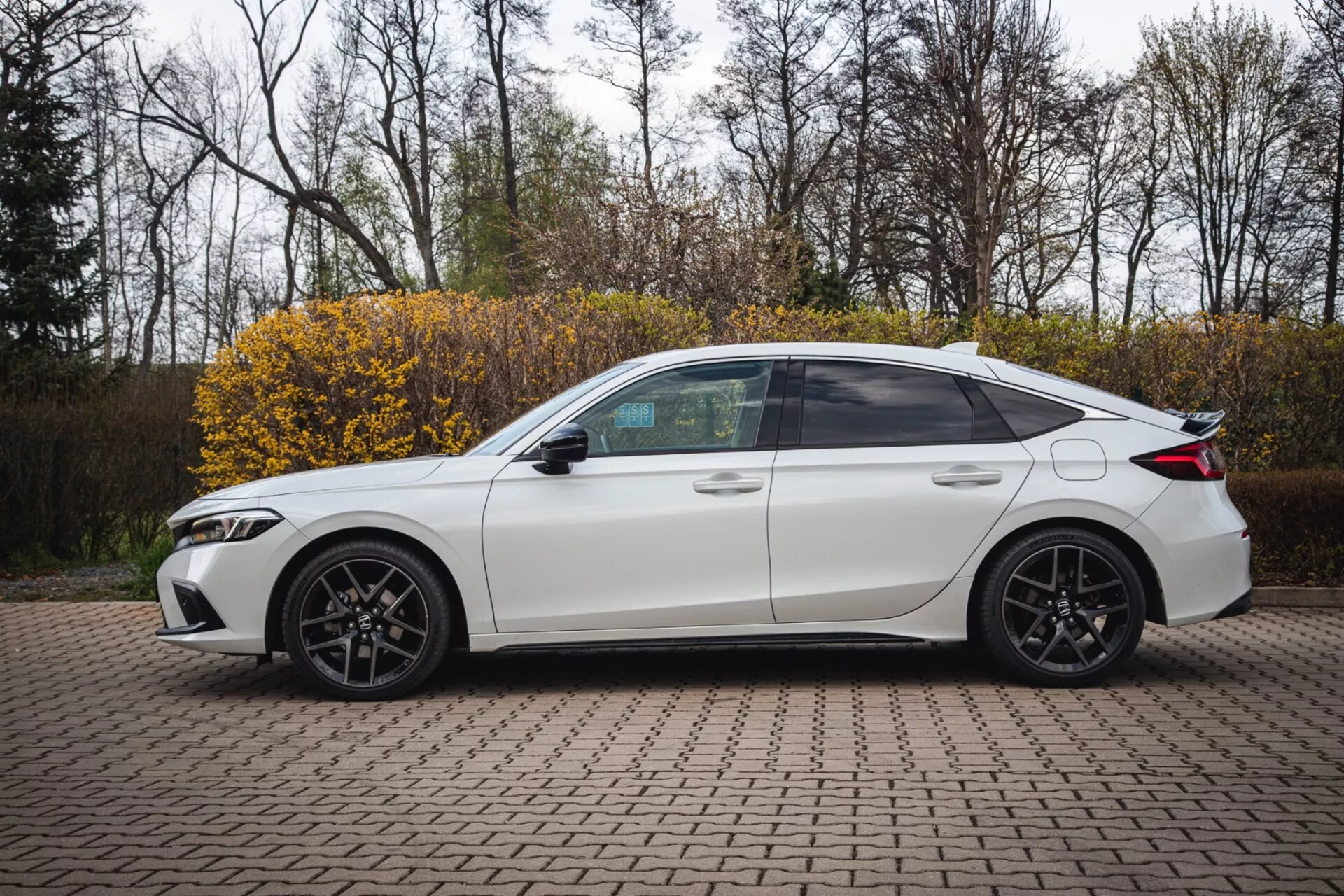 Beli štirivratni sedan Honda Civic letnik 2022, parkiran na tlakovani površini z zaklepanimi opekami, prikazuje stranski profil s športnim dizajnom in sodobnimi LED žarometi, posnet iz rahlo dvignjenega kota za poudarjanje celotnega stranskega pogleda vključno s temnejšimi okni, športnimi zlitimi kolesi in tehnološkimi značilnostmi, kot so vgrajeni smerokazi v stranska ogledala.