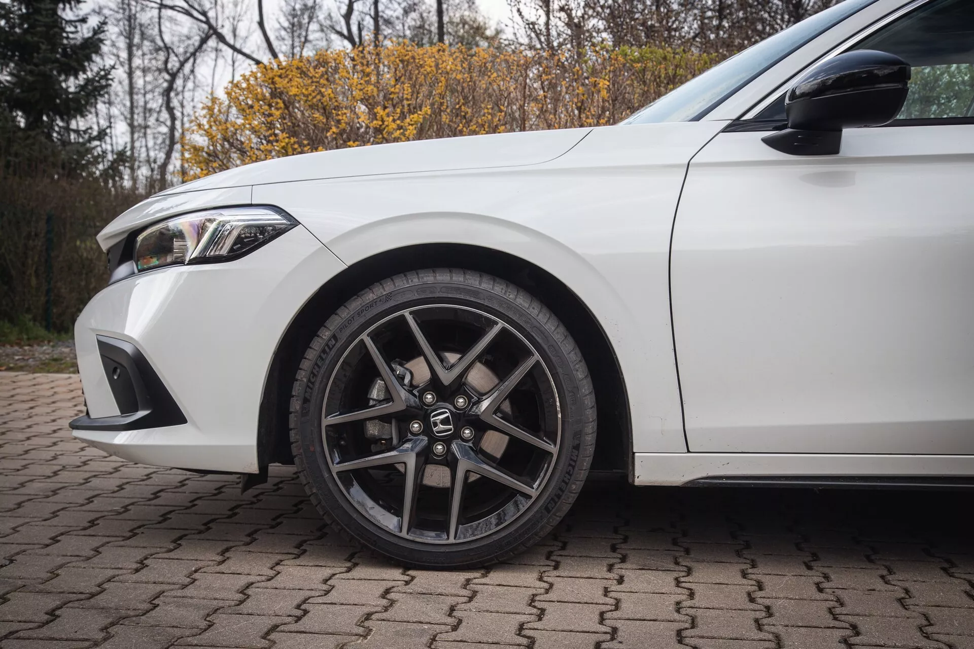Beli Honda Civic 2022, prikazuje sprednje levo kolo z večkrako zlitinsko zasnovo in pnevmatiko z nizkim profilom ter podrobnosti blatnika in dela sprednjega odbijača na opečnati površini