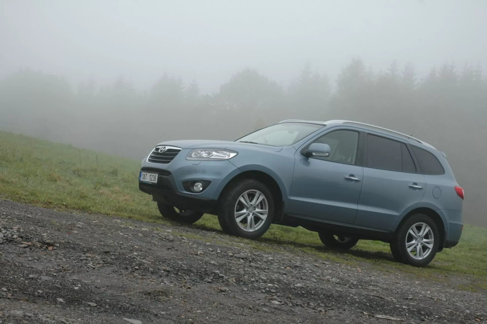 Modro srednje velik crossover SUV Hyundai Santa Fe letnik 2006 na neasfaltirani cesti z obližjem trave, zorni kot spredaj levo v meglenem vremenu poudarja robusten dizajn, sposobnost vožnje izven cest in sodobne lastnosti družinskega vozila, kot so sprednja rešetka, kotni žarometi, meglenke, lita kolesa, strešne letve in vgrajeni smerniki.