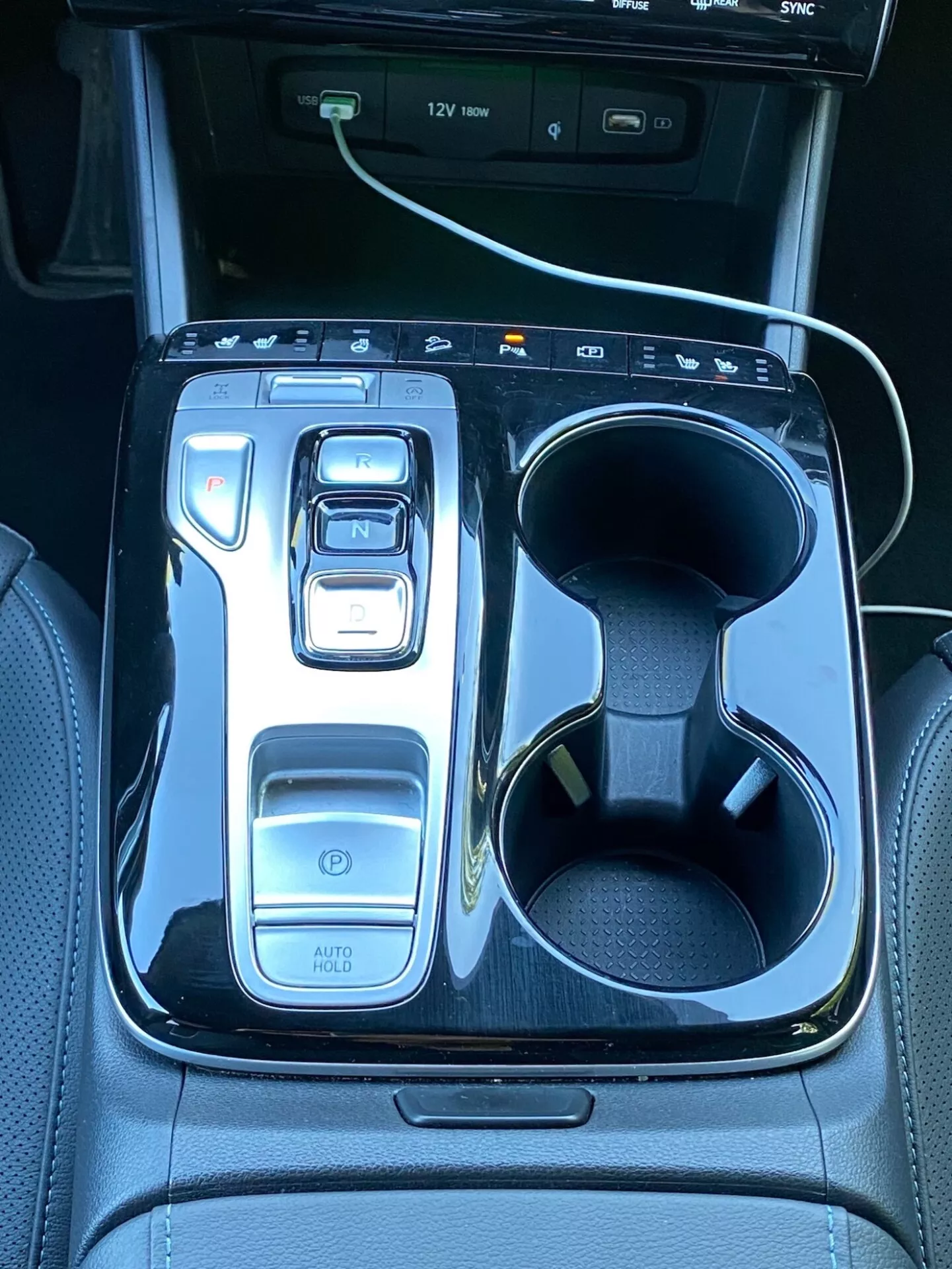 Slika prikazuje sredinsko konzolo Hyundai Tucson 2020, z elektronskim prestavnim vzvodom v položaju 'N', obkroženim z bleščečo črno oblogo. Na levi strani so kontrole, vključno z gumbom za napajanje, izbirniki načinov vožnje in funkcijo 'AUTO HOLD'. Na desni sta dva nosilca za pijačo, eden z belim polnilnim kablom. V zgornjem delu je viden del zaslona za info-zabavo z USB priključki, kar poudarja tehnologijo in luksuz vozila.
